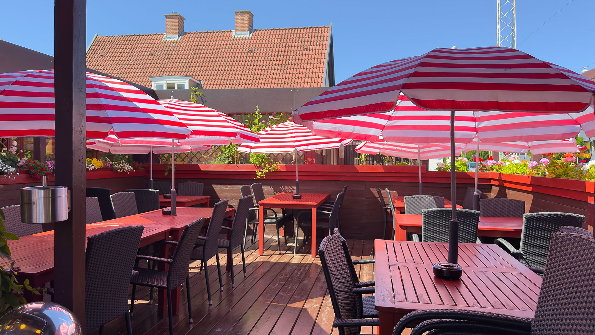 Terrasse med røde og hvide parasoller og stole, hvor der er blomster og et rødt hus i baggrunden