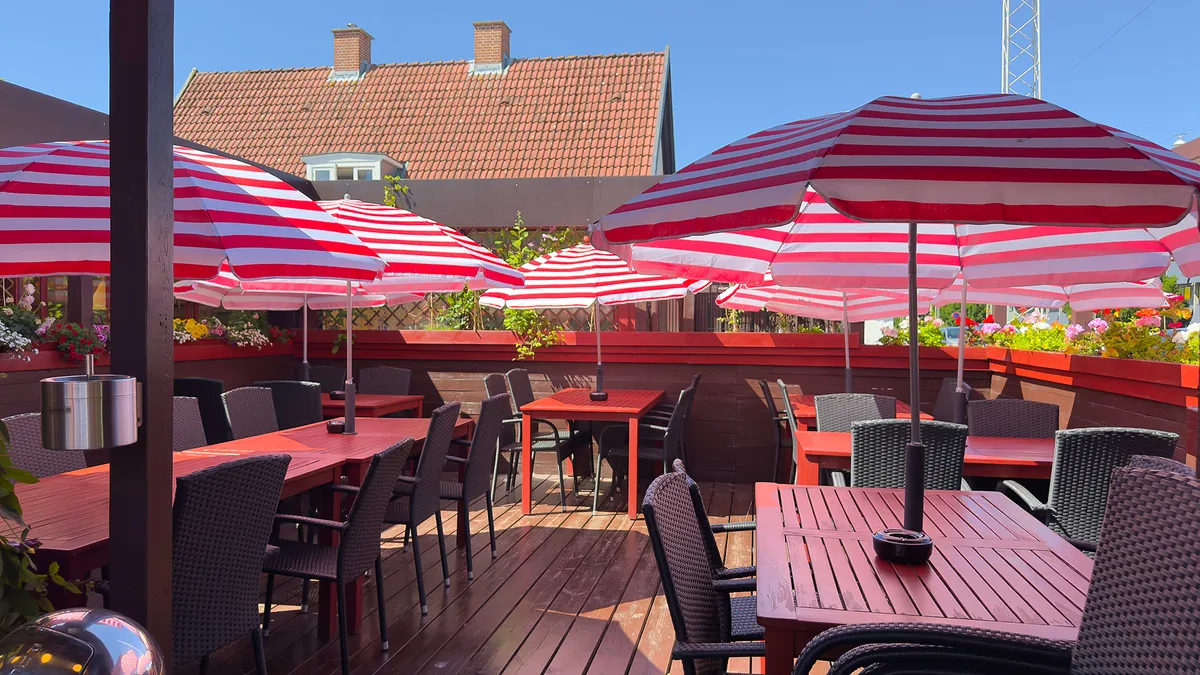 Terrasse med røde og hvide parasoller og stole, hvor der er blomster og et rødt hus i baggrunden