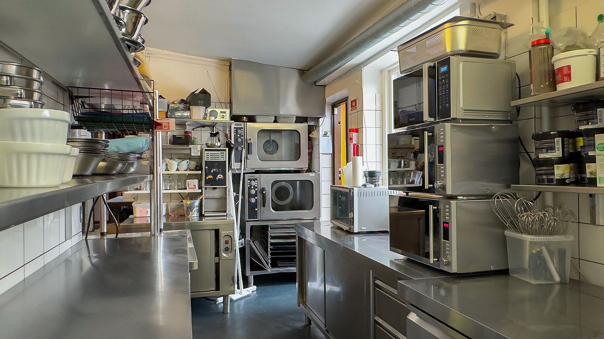 Et luksuriøst køkken med moderne apparater, kedler, og en lang metalarbejdsbord. Der er en række mikroovne, kogebøtter, og
