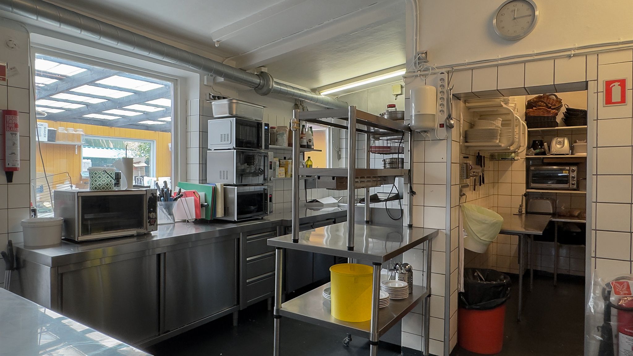 Et køkken med flere mikrobølgeovne og en kaffemaskine. En metalvogn med flere hylder og et gult fad. En gulvstøvsuger og en