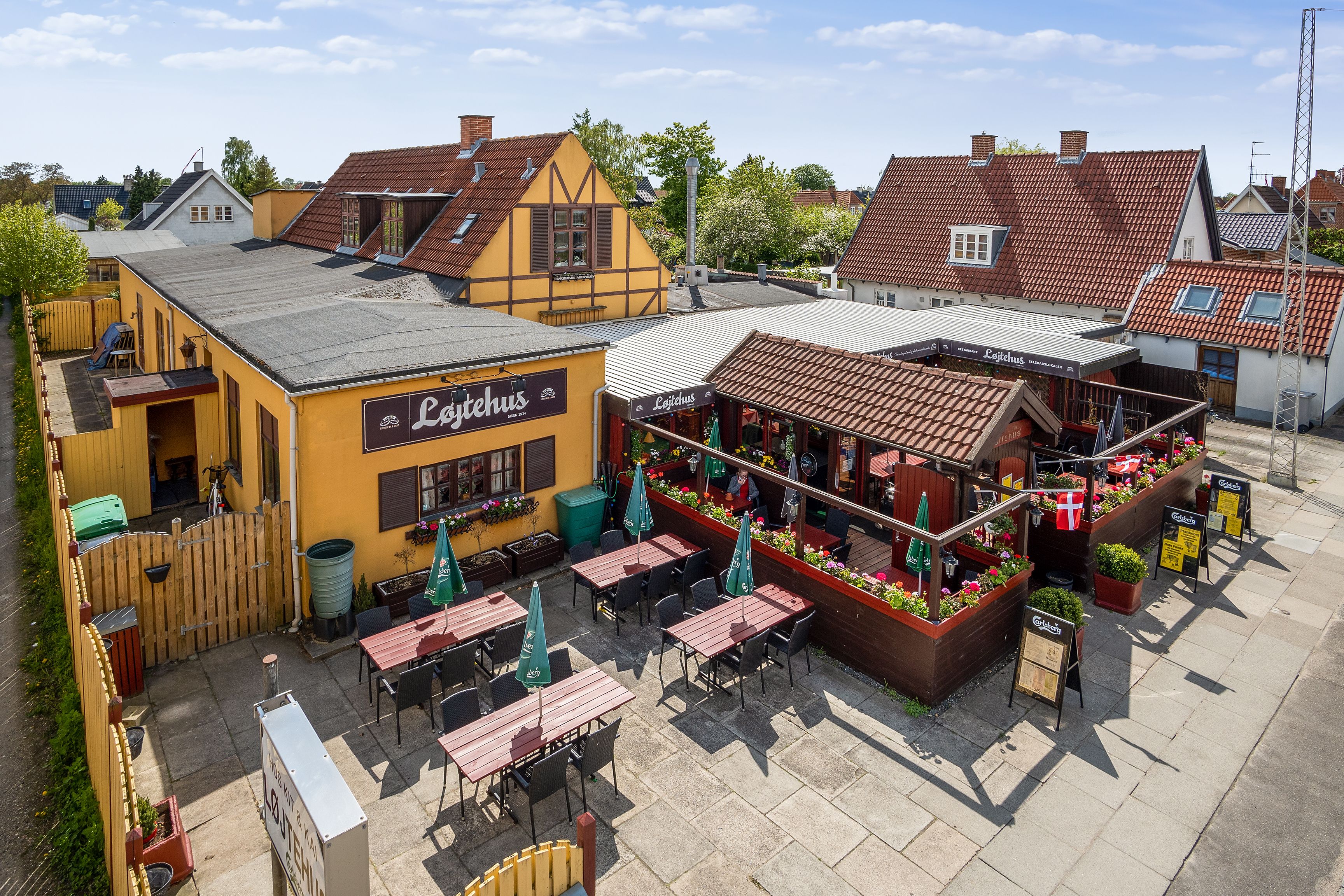 Restaurant med udendørs siddepladser og grønne parasoller samt røde og gule bygninger med røde tag og mange blomster.