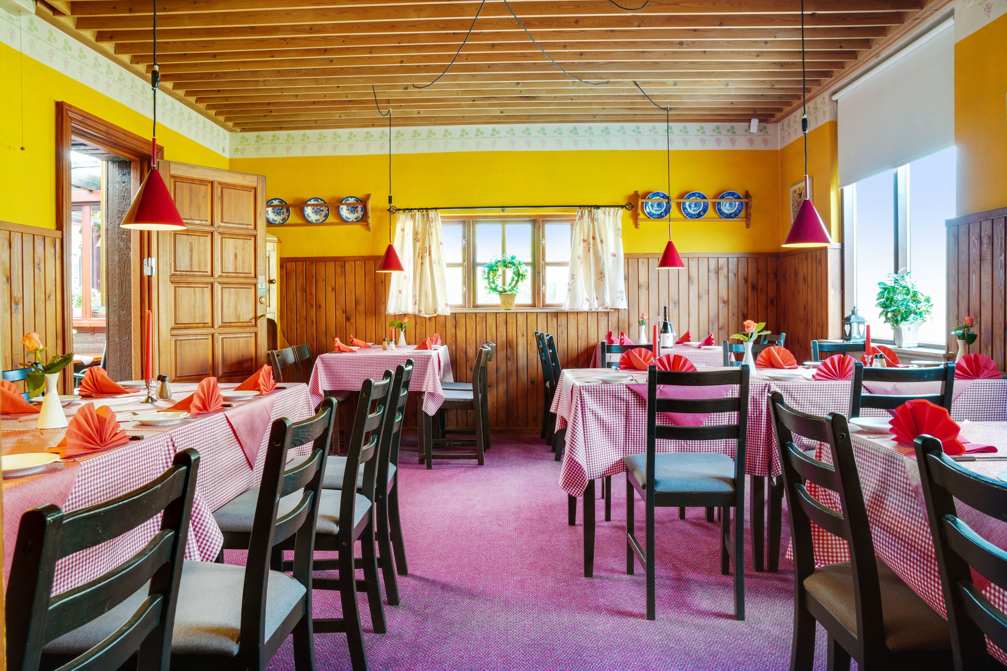 Bord med røde og hvide kuverter, og røde lampeskifter i en restaurant med en gul væg og en træloft