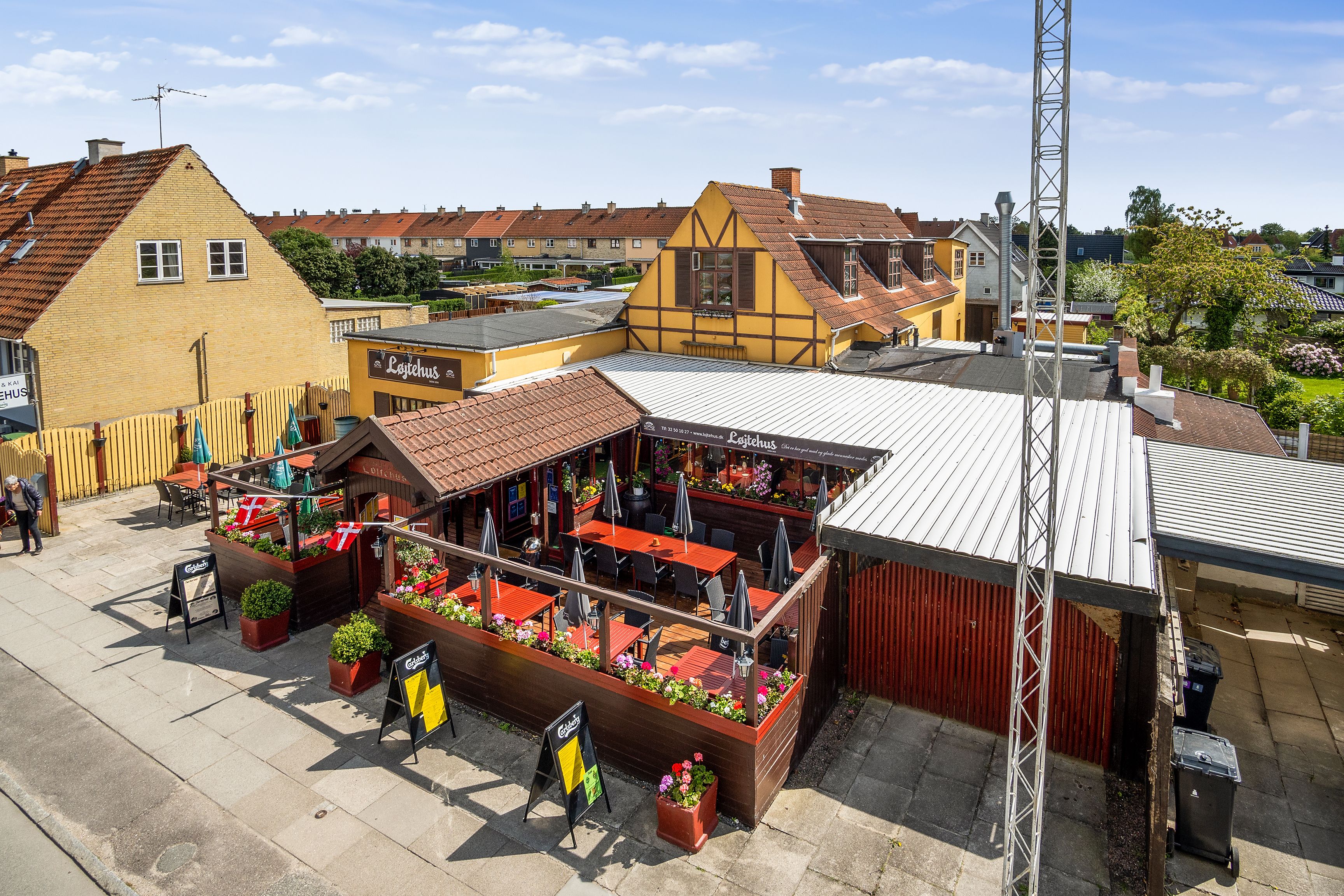 Overblik af Løjtegårds restaurant i Kastrup med terrasse og bord på forsiden. Røde og gule bygninger med blomsterpotter og