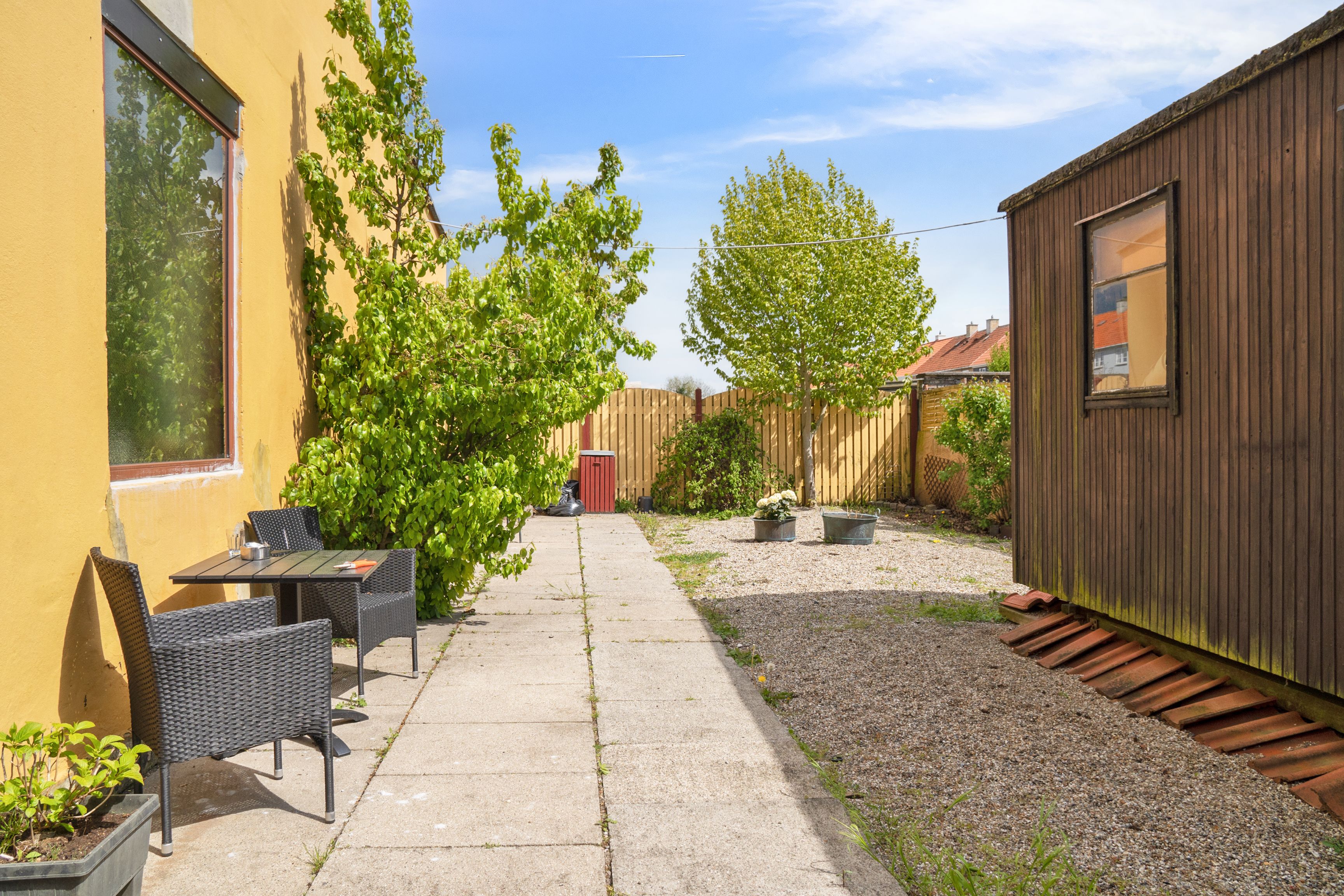 Afslåede gårdhave med gul væg, spiseplads med to stole, grønne planter og en lille huse i baggrunden med en grøn bakke