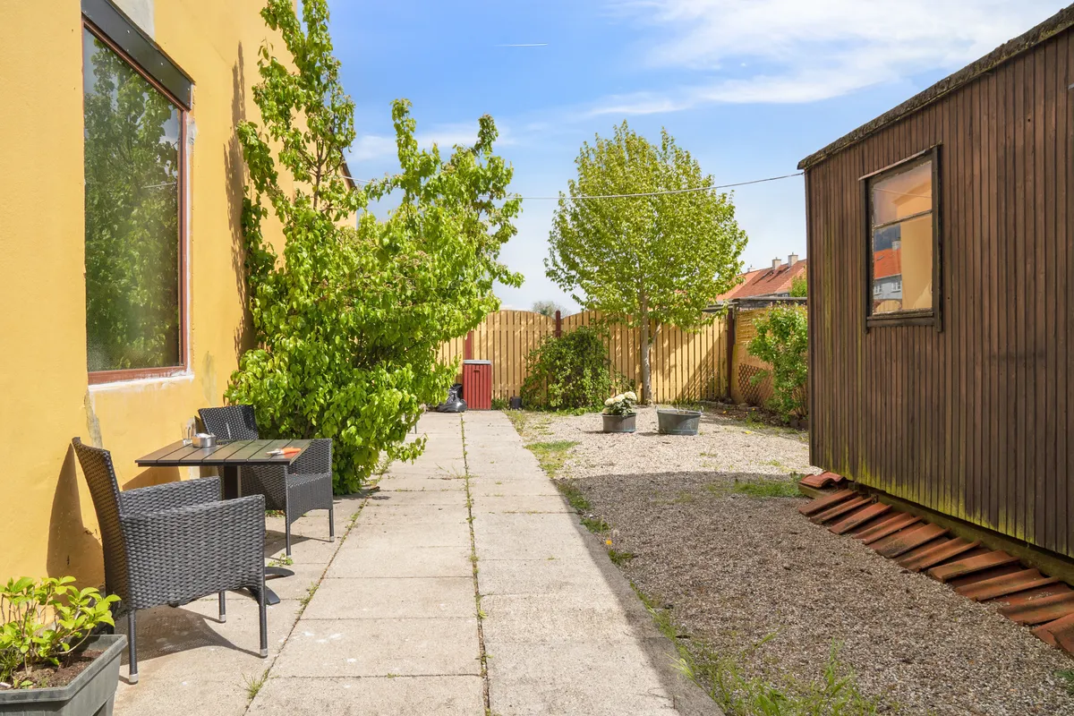 Afslåede gårdhave med gul væg, spiseplads med to stole, grønne planter og en lille huse i baggrunden med en grøn bakke