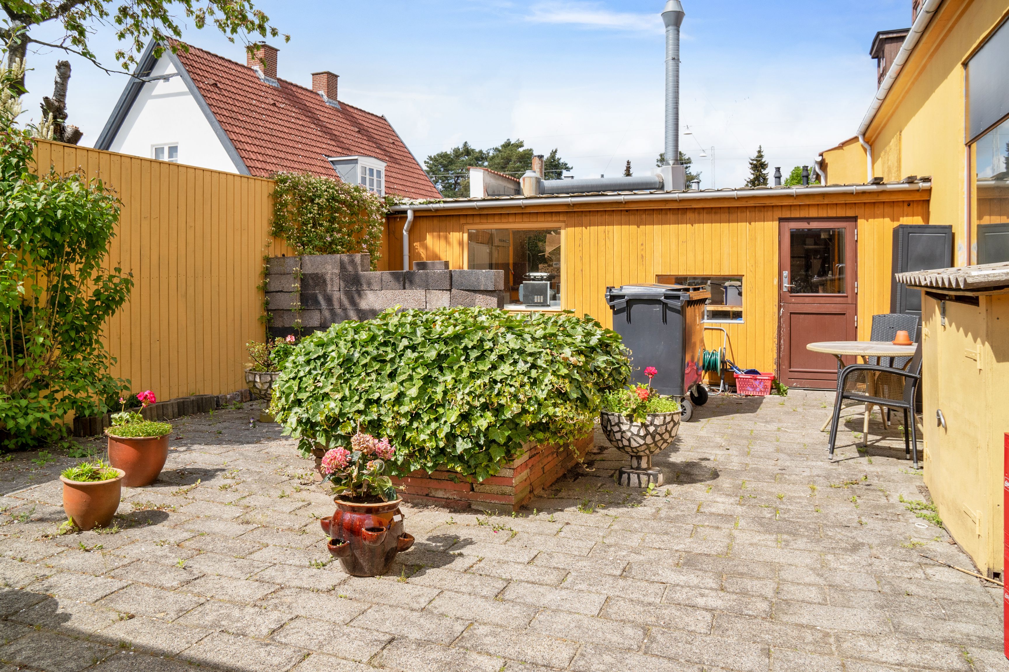 Have med blomsterplanter og gul skur i baggrunden. Bord og stol med krukke samt en større skraldspande.