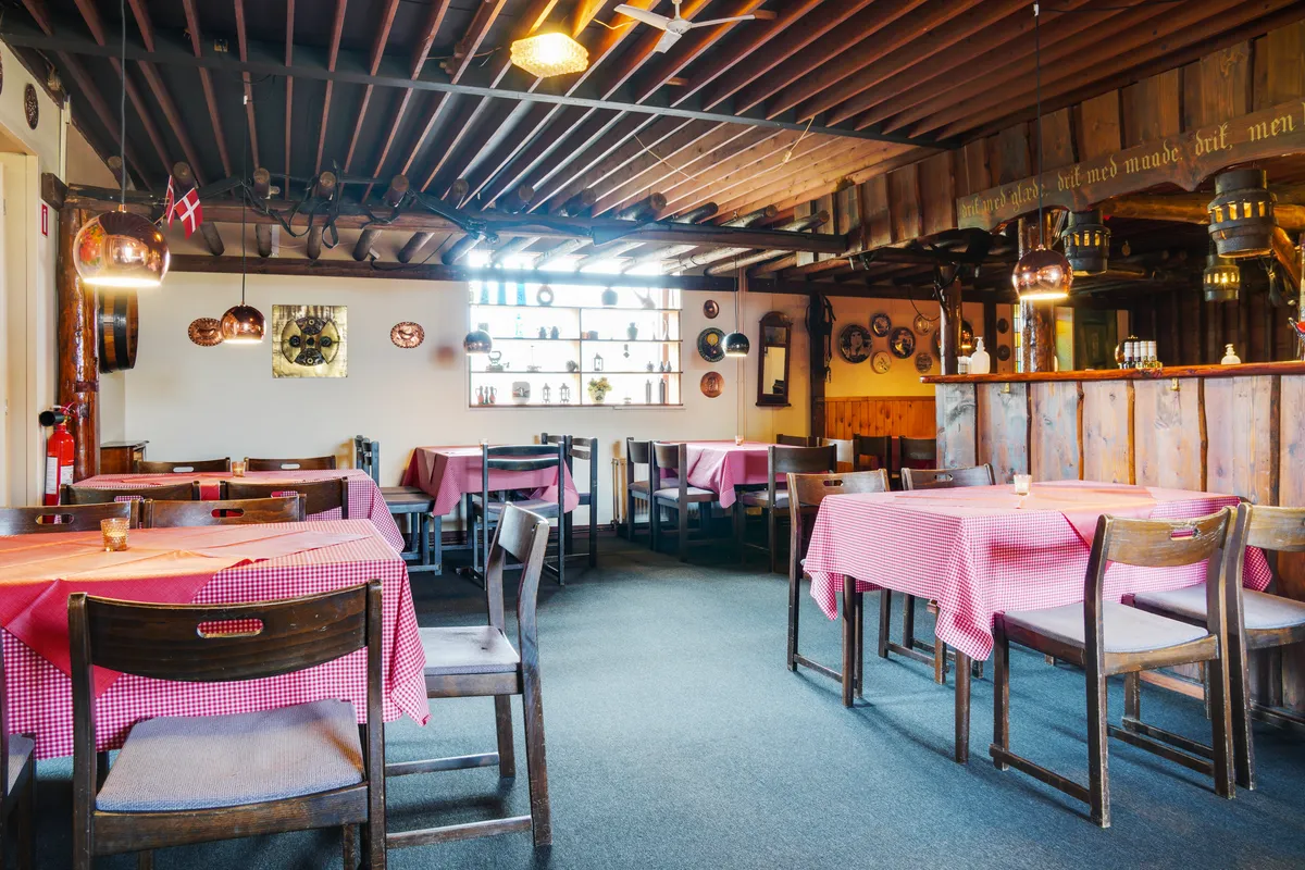 En restaurant med røde og hvide borddækninger og lyse træstole og stolpe. Væggene er lavet af træ og dekoreret med en række
