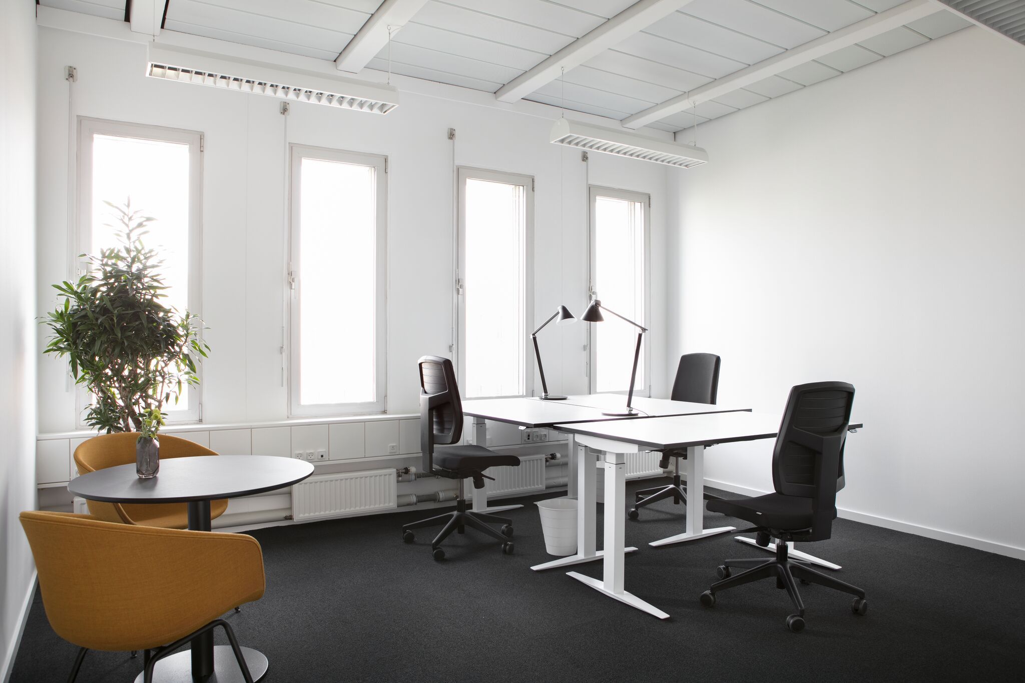 Det er et kontor med fire skriveborde, to høje bord med lampe og to lavere. Alle bord er hvide med sort stof. Der er fire