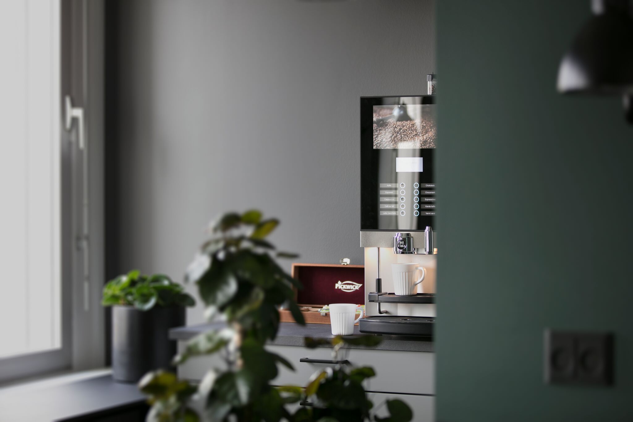 Kaffemaskine står på kontorbord med to krus, plante og en mørk væg med vinduesvisning.