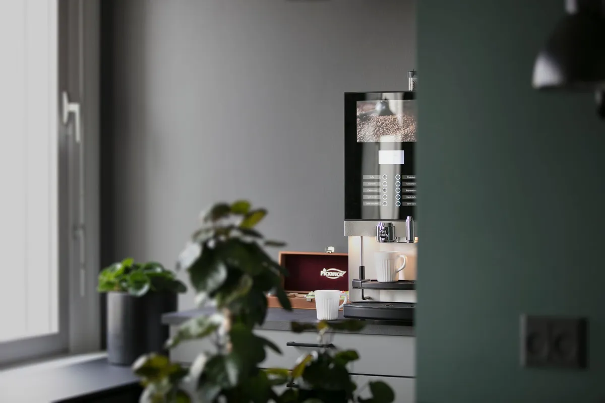 Kaffemaskine står på kontorbord med to krus, plante og en mørk væg med vinduesvisning.