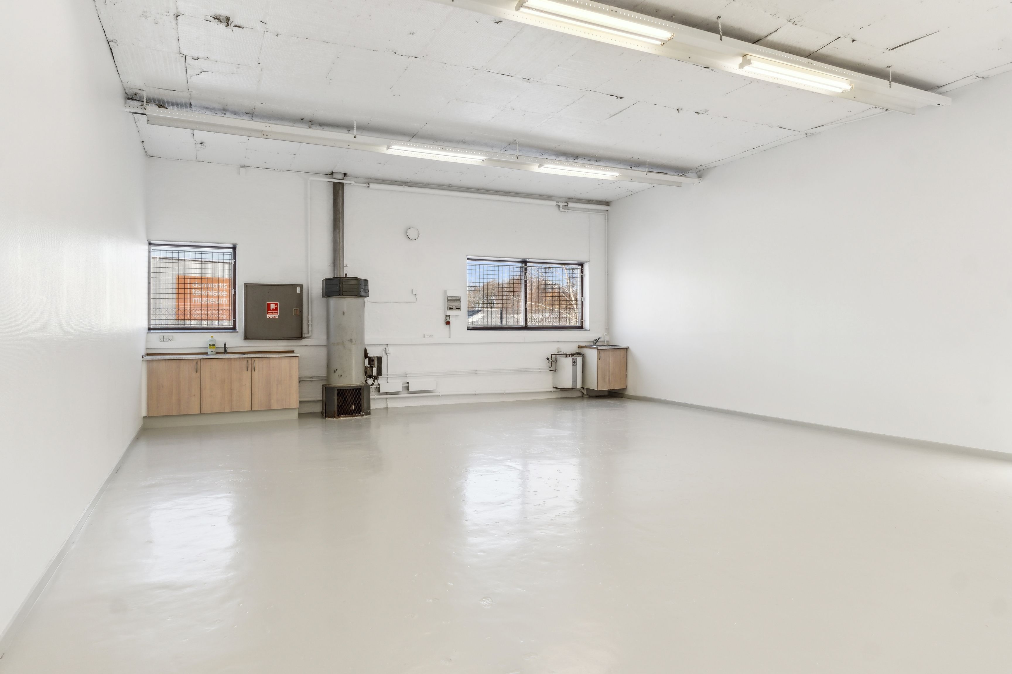 Et rum med hvide vægge og loft, et vindue med lysegardin, et skab og et toilet på højre side samt en ovn og et varmeapparat