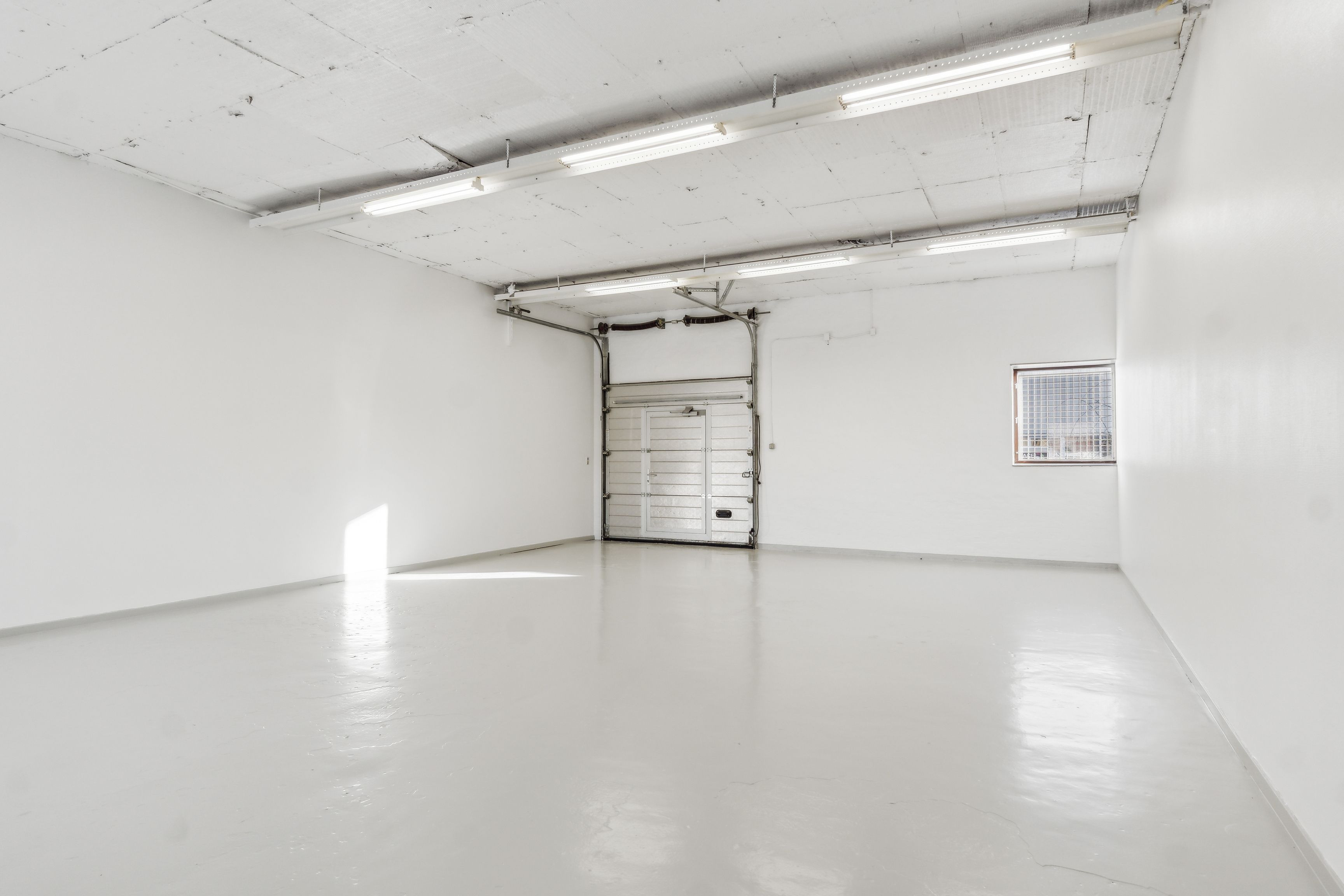 En stor, ubenyttet erhvervslokal med lysegulv og hvide vægge samt en garagedør og et lille vindue