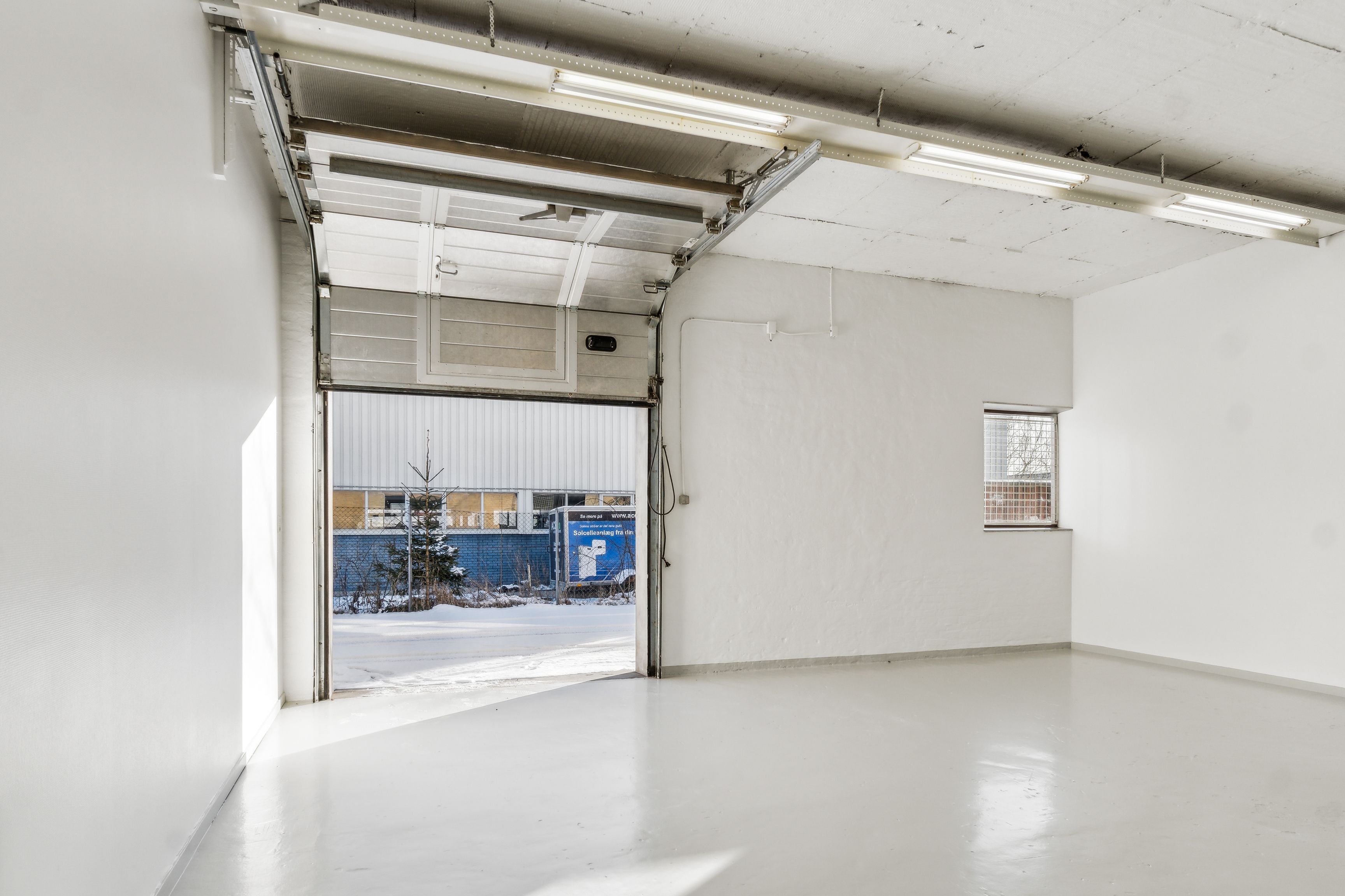 Et ubenyttet og hvidmalet lokale med åben garagedør og indgang til en gård med sne, der er omgivet af en hvidmur og en