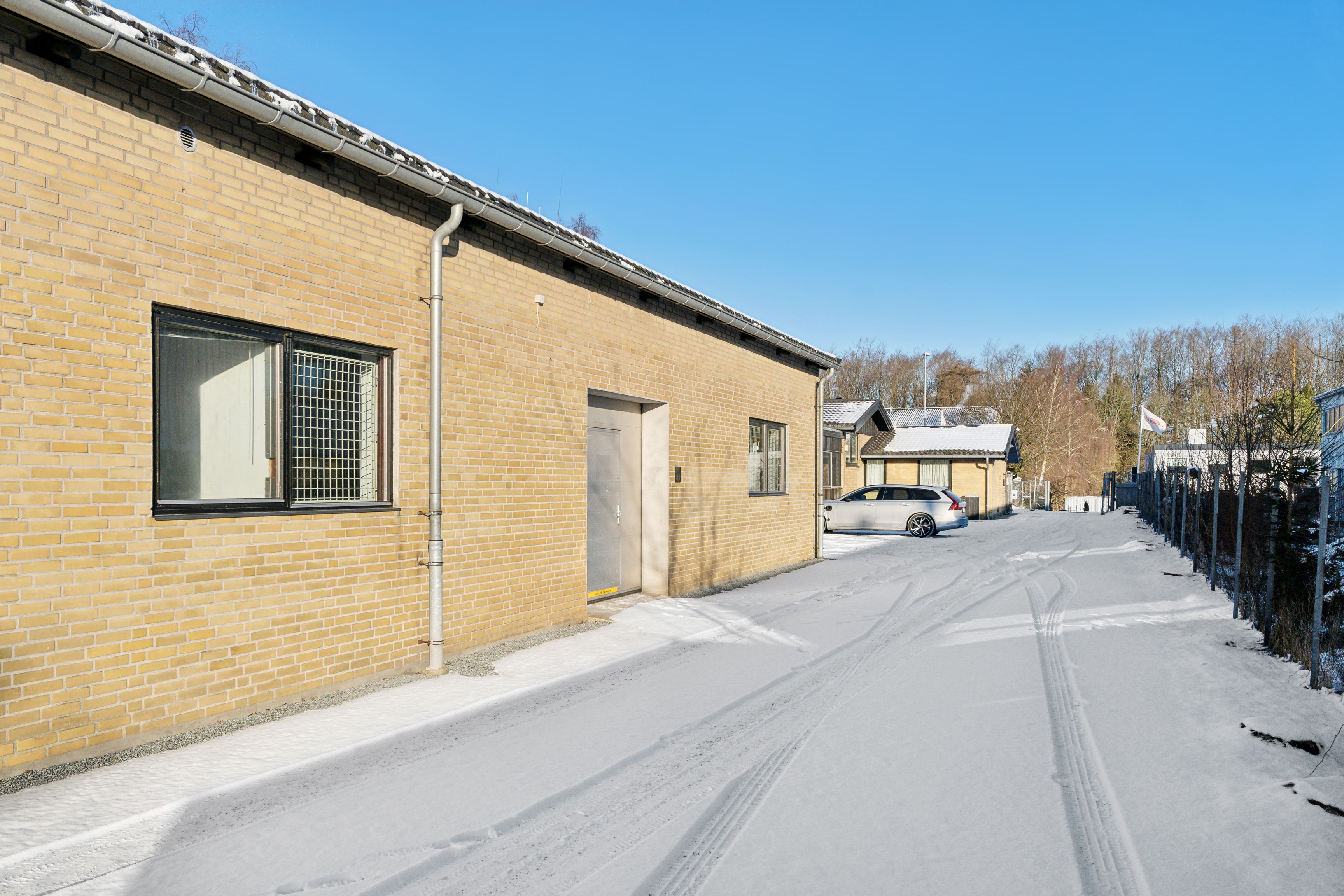 En hvid bil parkeret ved en garage i en snebelagt vej, med en gule murstensbygning og en flagpole i baggrunden