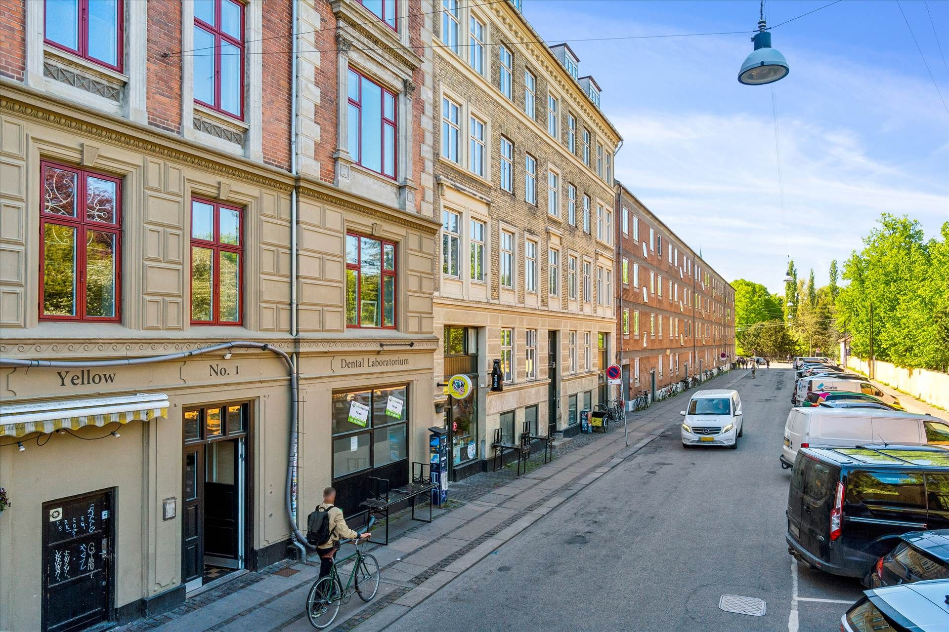 Man cykler på en gågade med flere biler parkeret langs siderne, og foran står en mand med en cykel. Der er en gågade med en
