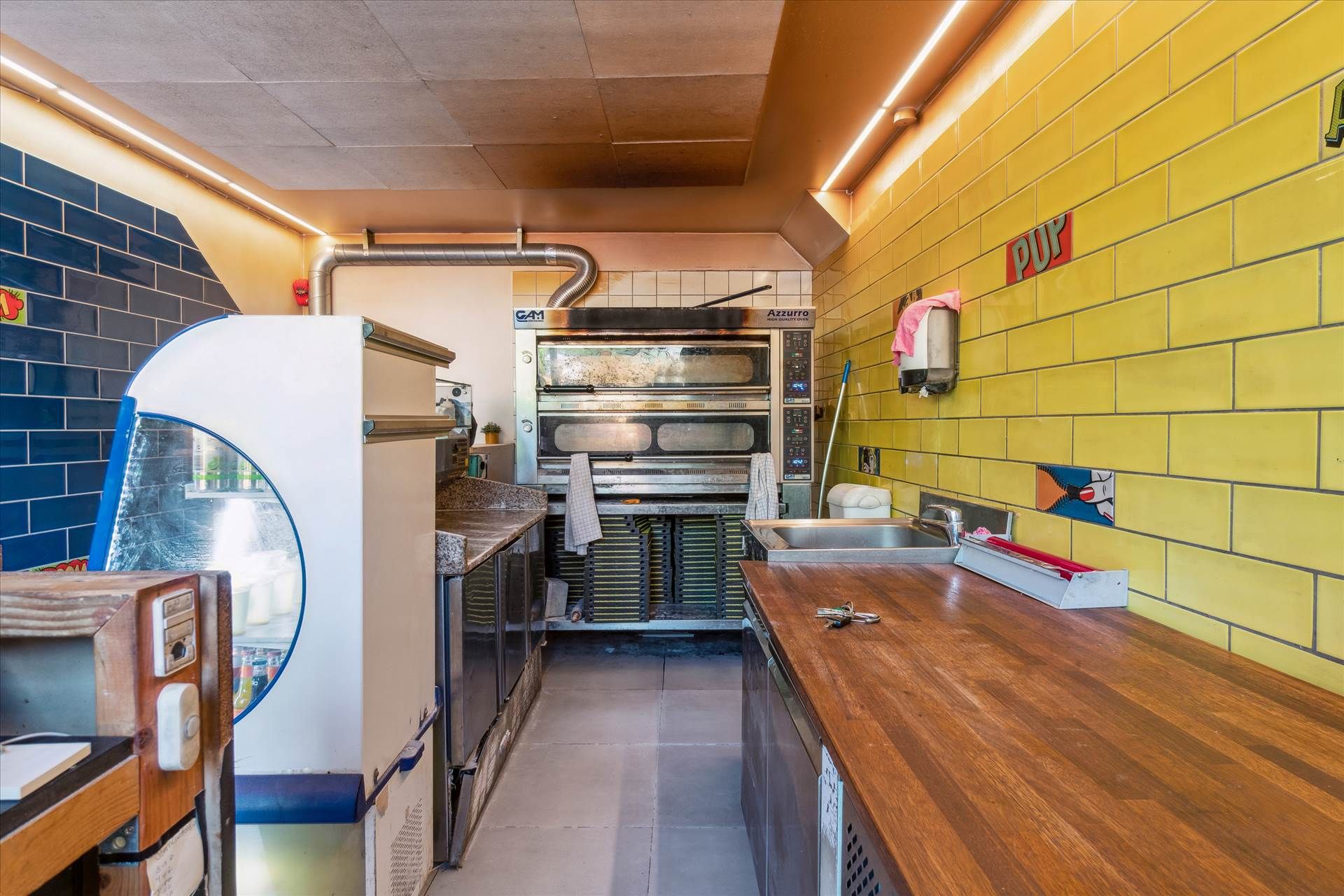 Køkken med træbord, slibemaskine, ovn og væg med blå og gule fliser samt et papirhåndklæde i holderen