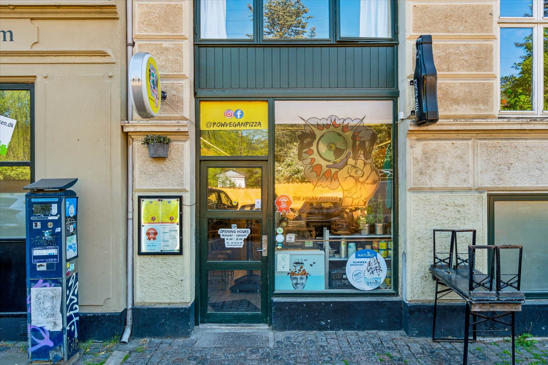Vegan pizzarestaurant med vinduer og dør i grønt og gulefarvet glas og en terrasse med stole på forsiden