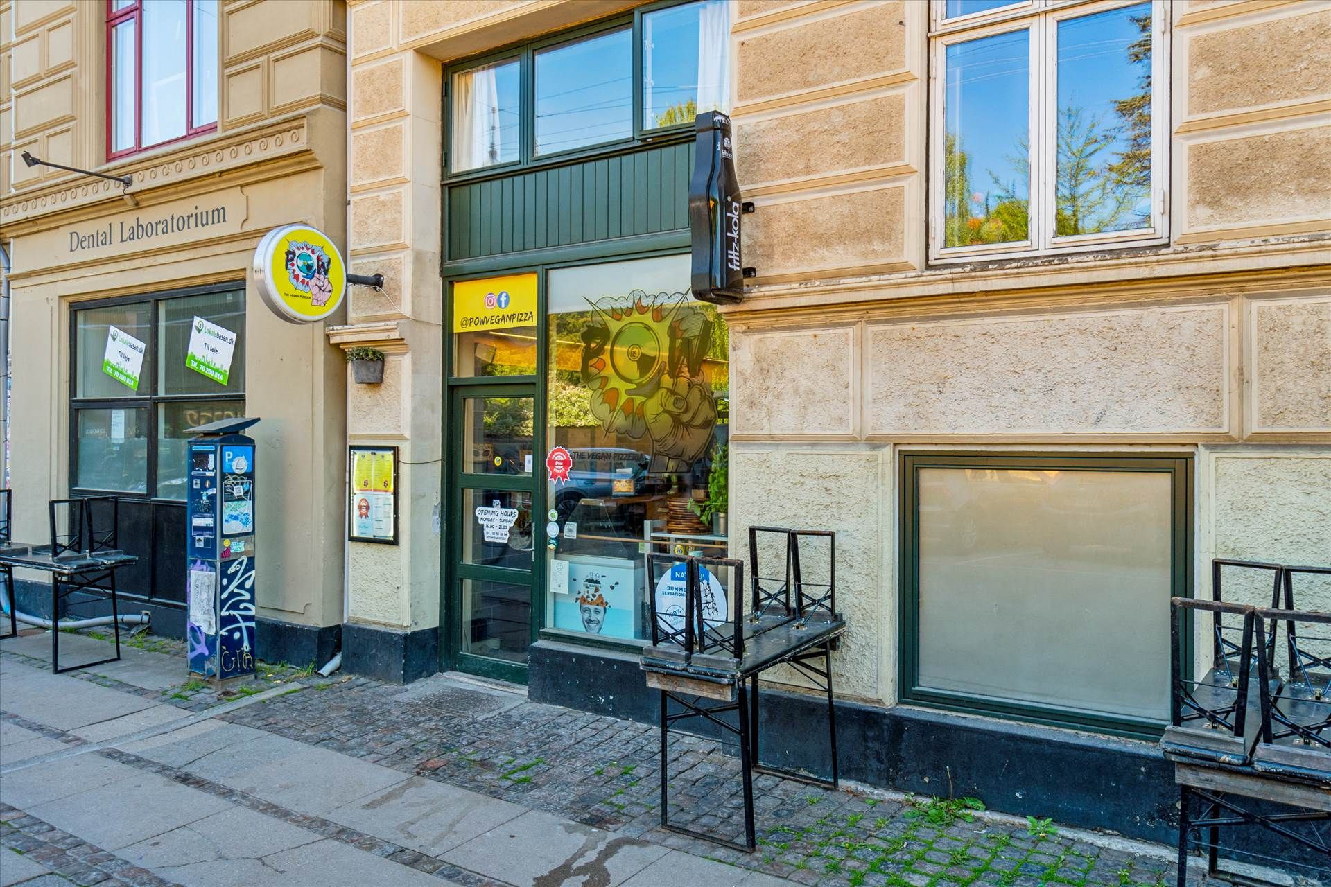 Forretningslokaler med åbent glasdør og træstole udenfor på Nørrebro. En sodavendingsautomat står på gågaden med skilte og