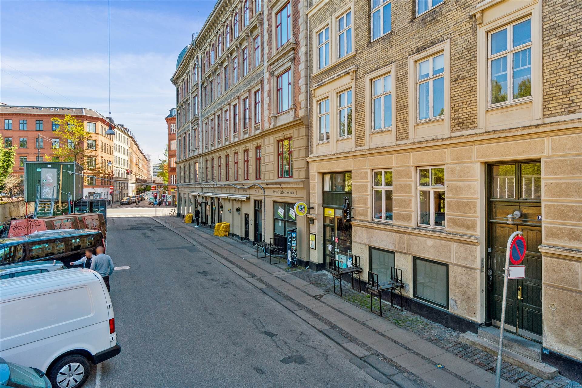 To parkede hvide lastbiler og en bil står på gaden med stribet fortov. To mænd står ved en hvid lastbil og snakker.