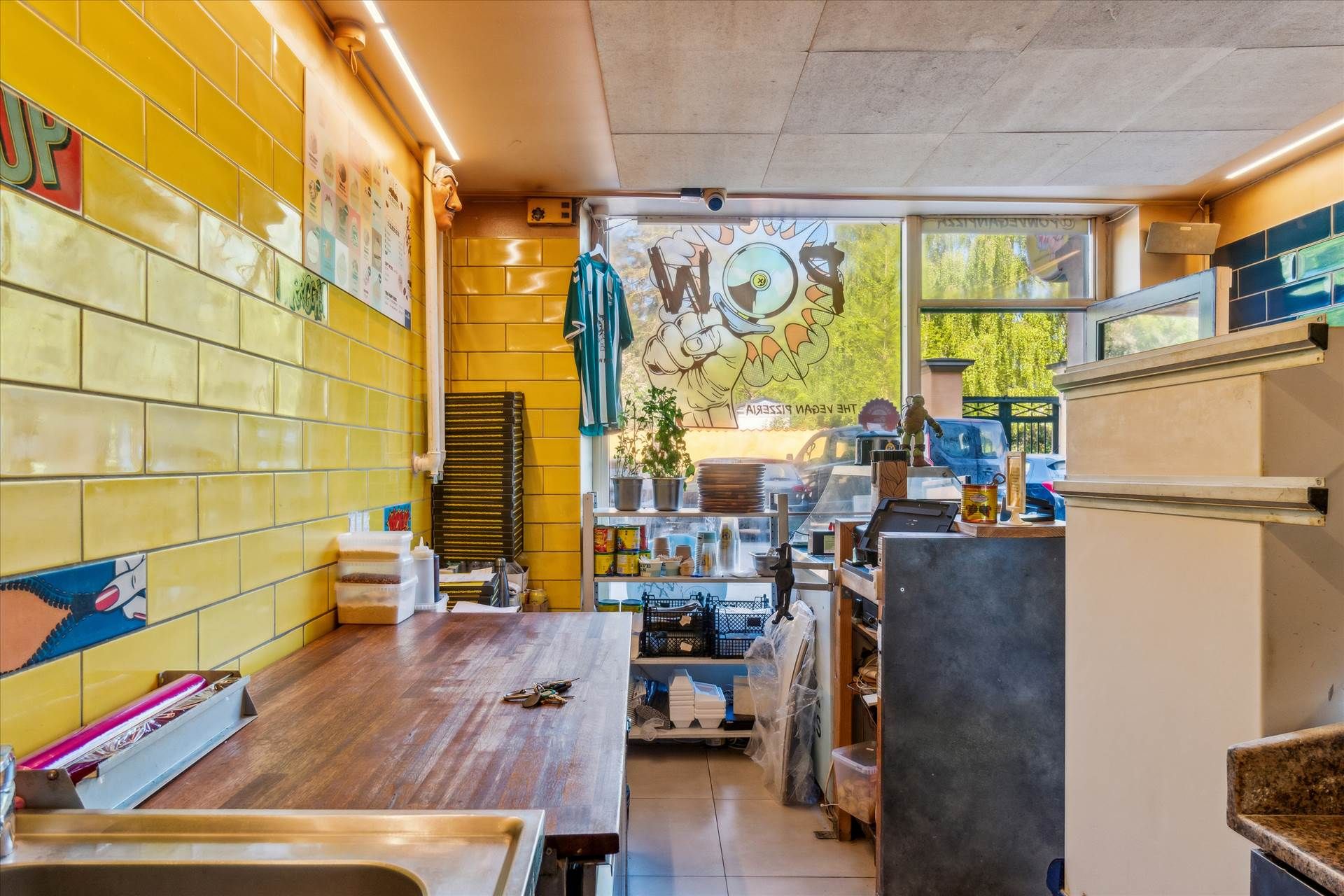 Køkken med lysegulle vægge, brædder, et bord, et køleskab, et sæt sager og en kæmpe skilt med "WOP" og en sol med briller