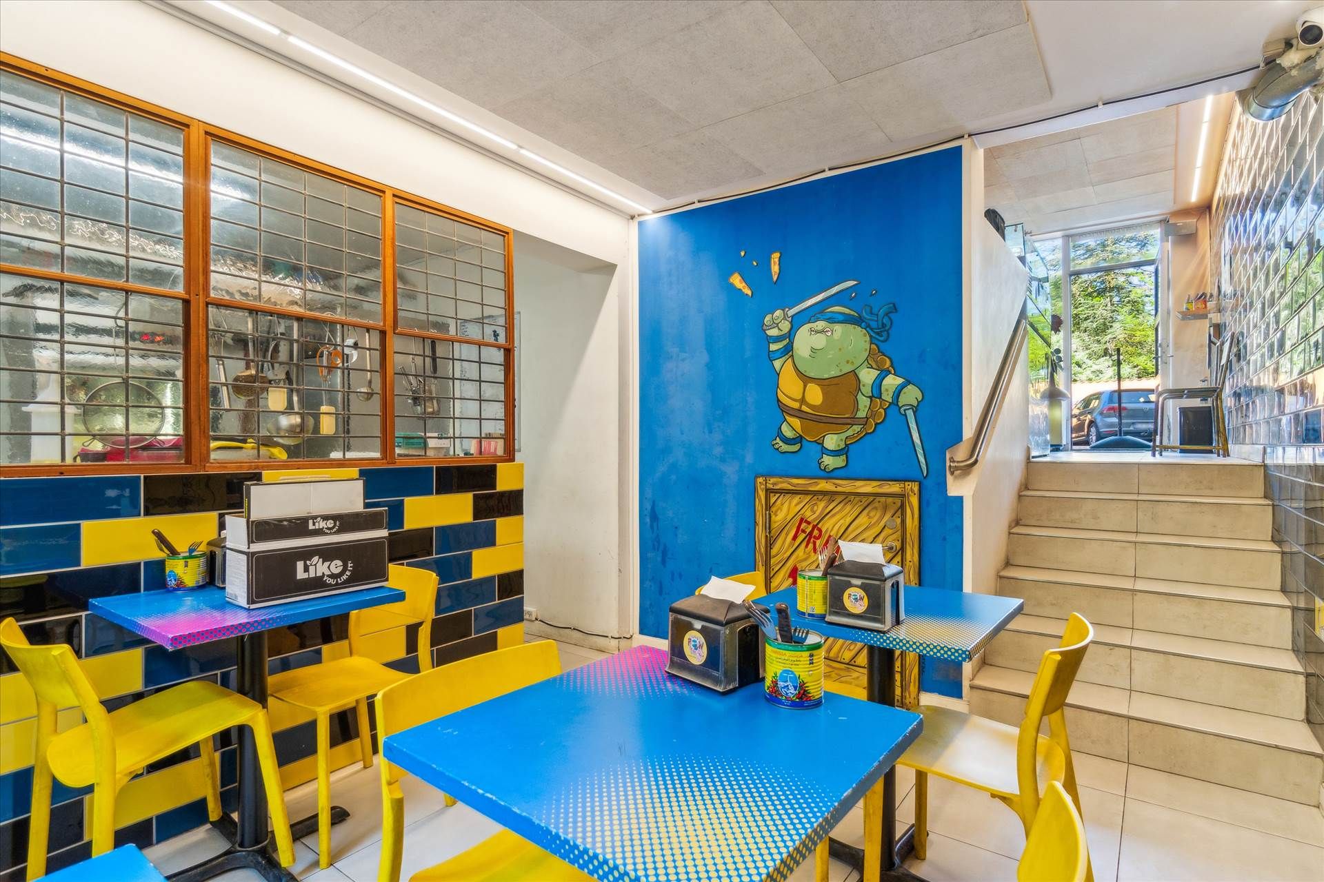 Gule stole og borde med krydsfiner og plast samt en væg med en tegnertegning af en ninjaskildpadde. Der er et skraldspand,