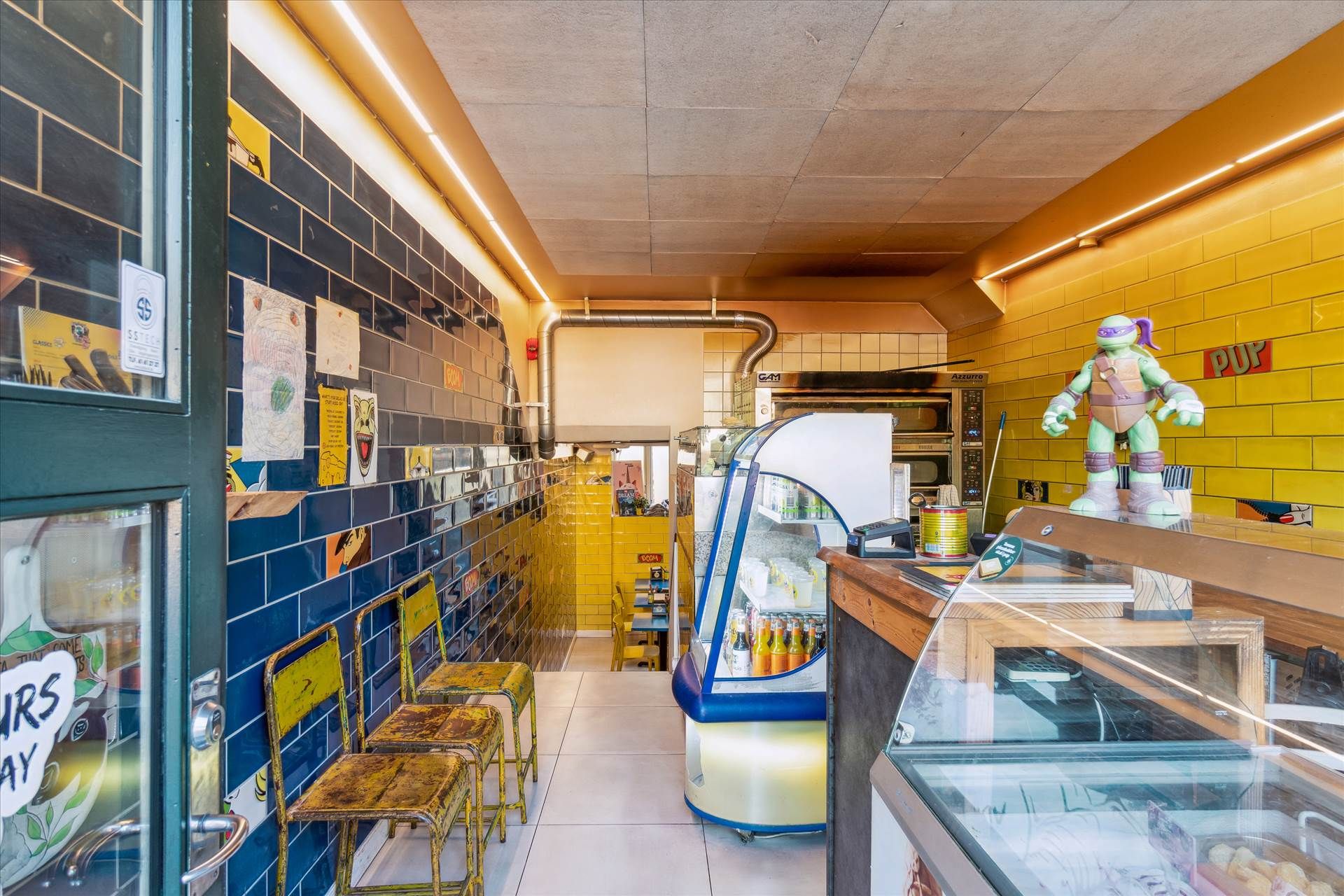 En lys og lækker madbutik med gul og orange interiør, en Ninja skjaldet på sælgeren og forskellige varer i udstilling