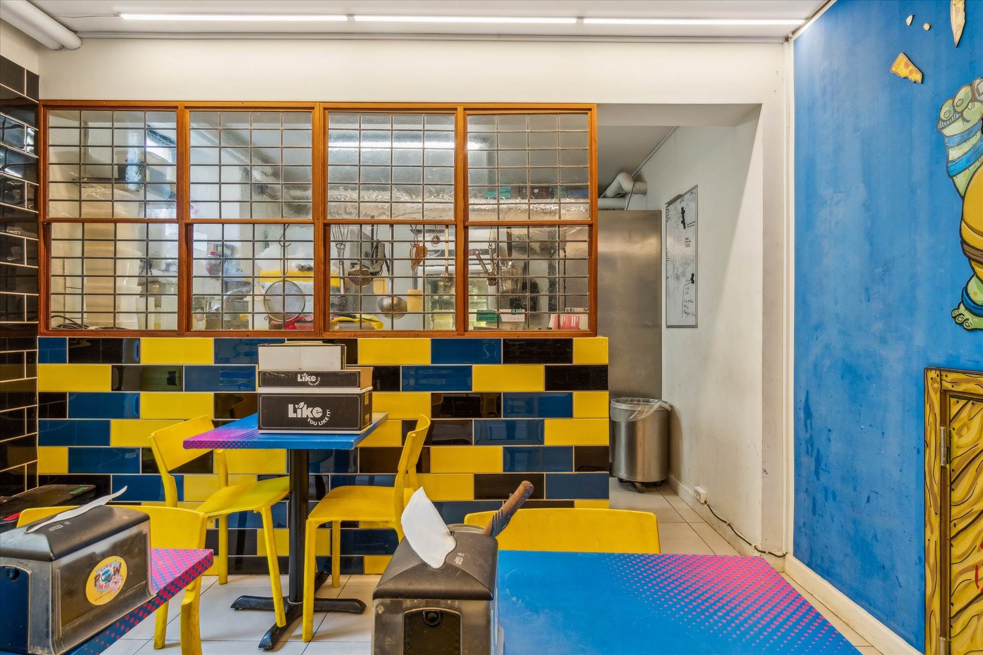 Køkken med gult og blåt tegl og lysegul væg, skrivebord med servietter og torskebøf, flere sædestole og vinduer med udsigt