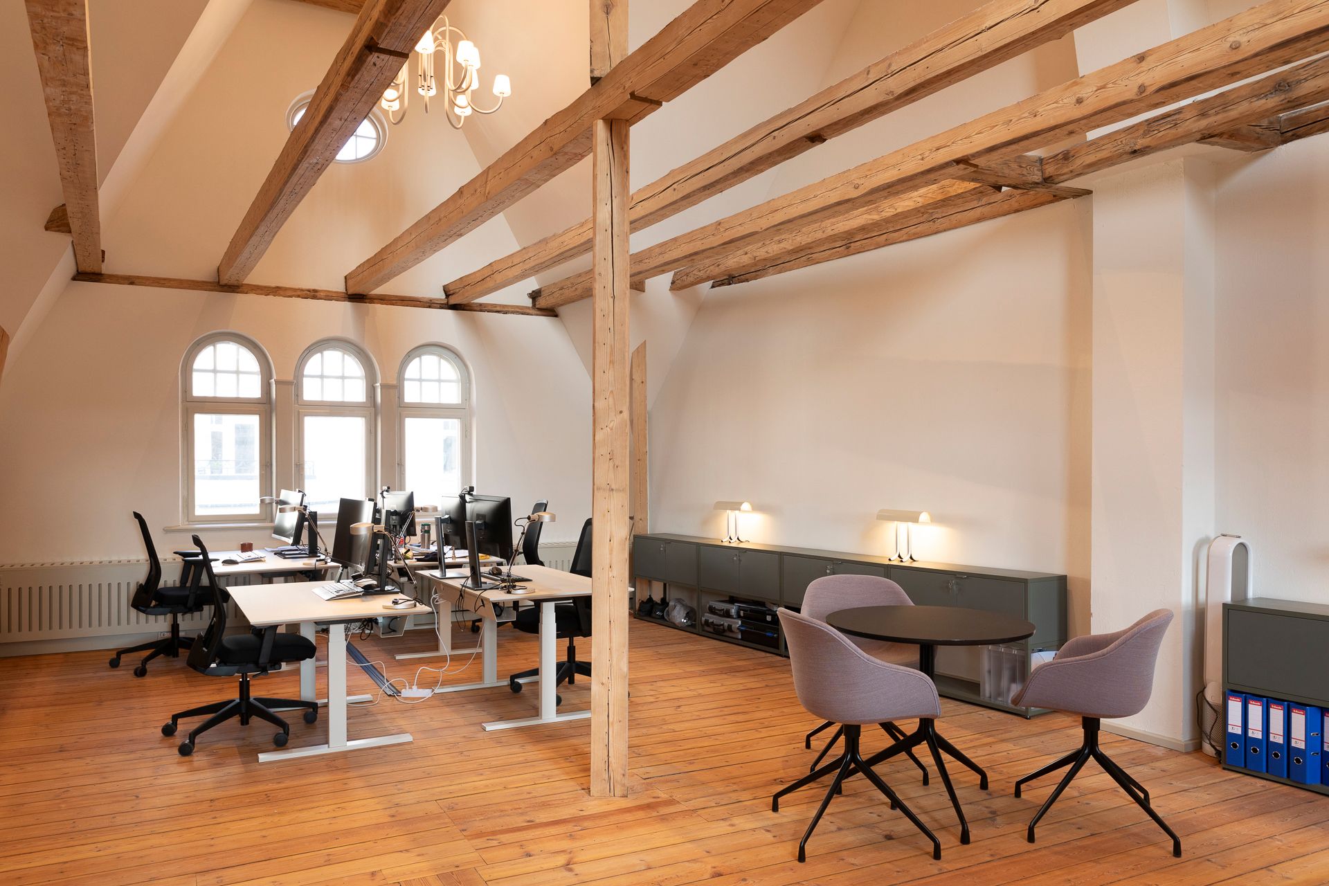 Kontor med lyse gulve og højt loft med lyse bjælker. Værelse med skriveborde, stole og computere. To borde med flere stole