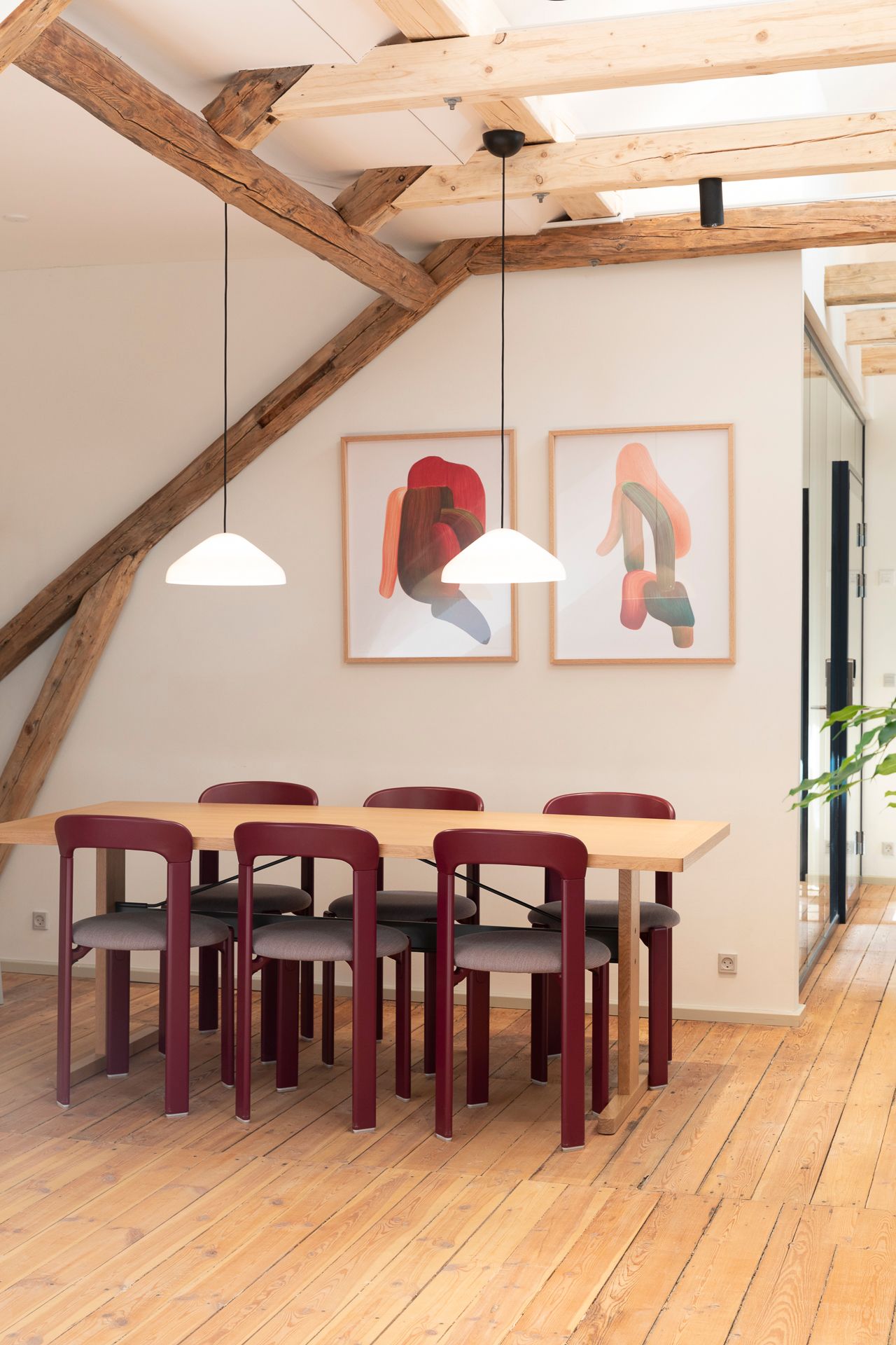 En spisebord med seks røde stole og to hængelampe er i et rum med højt loft og lyse loftbjælker.