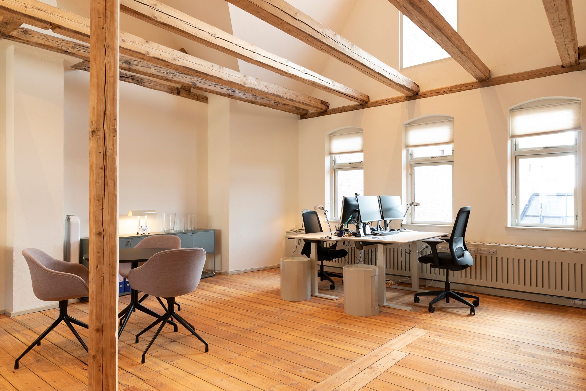 En stor og lys kontor med trægulv og træloft, flere skriveborde med computere, stolpe og møbler placeret rundt i rummet.