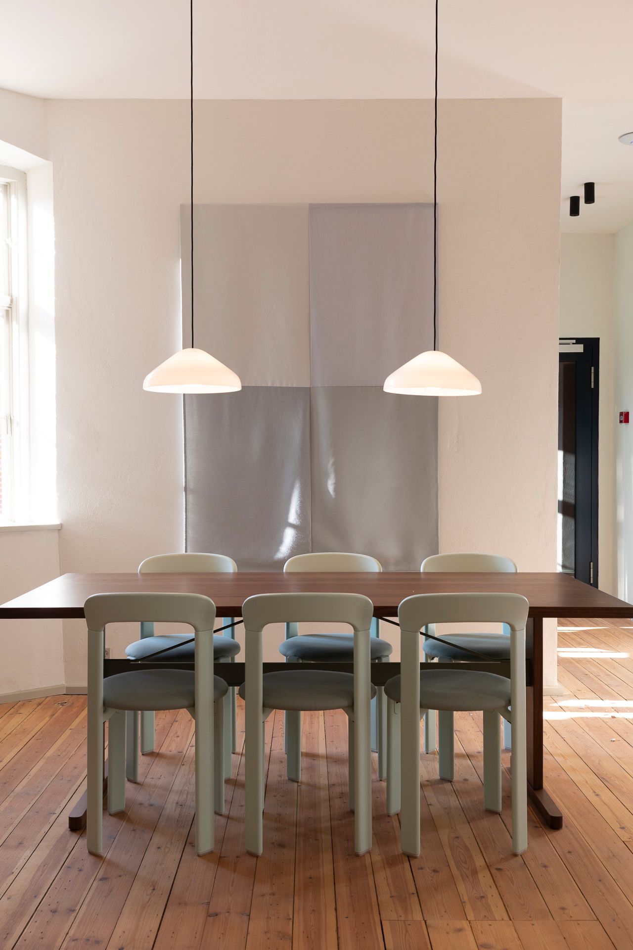Lys beige stole placeret omkring en mørkebrun bord med to hængelampe over bordet i et kontor med lyse vægge og trægulv