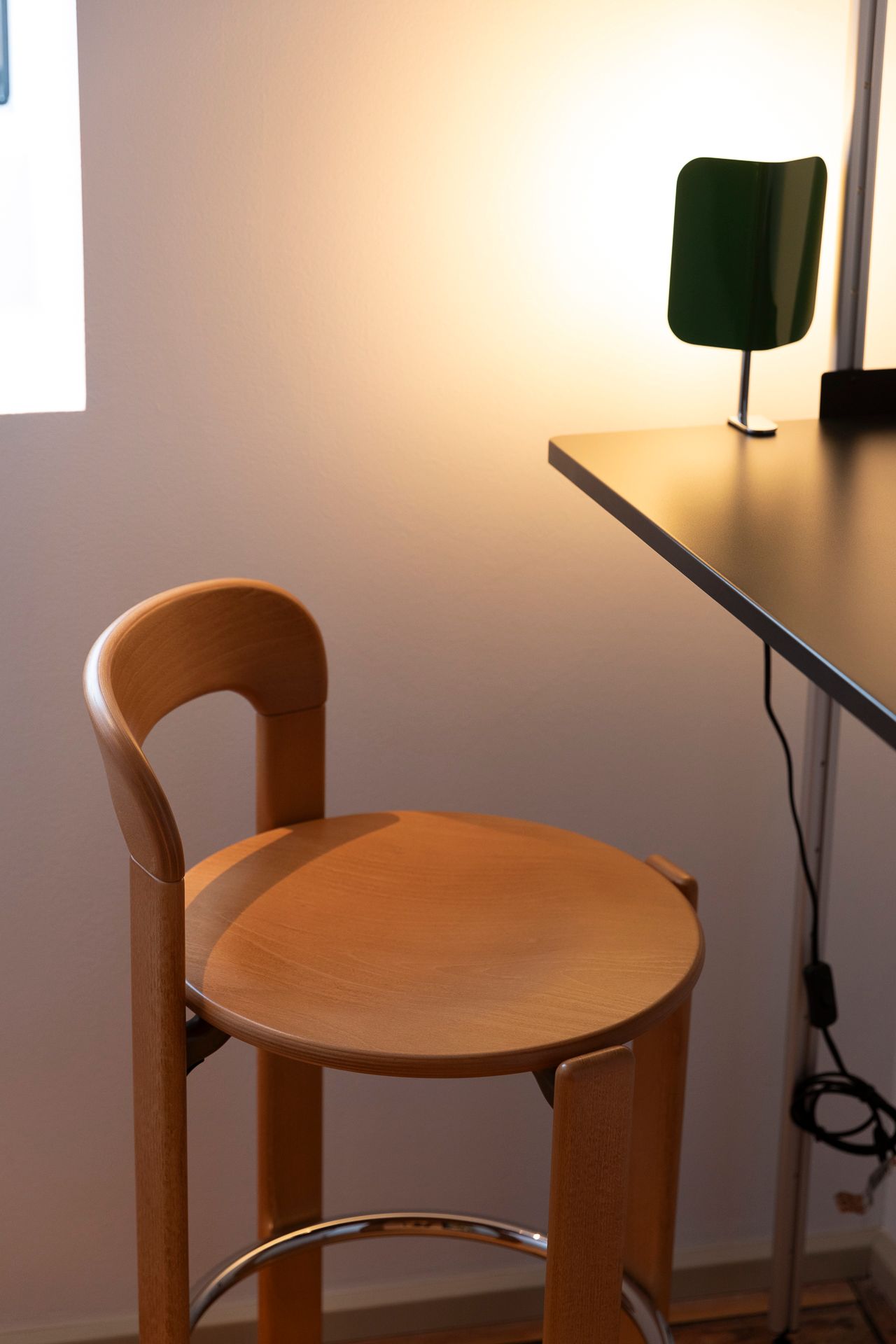 En lysegrå, moderne stol med skrå ryglæn står ud for en lys, lysegul væg og et langt, brunligt bord med en grøn, drejelig