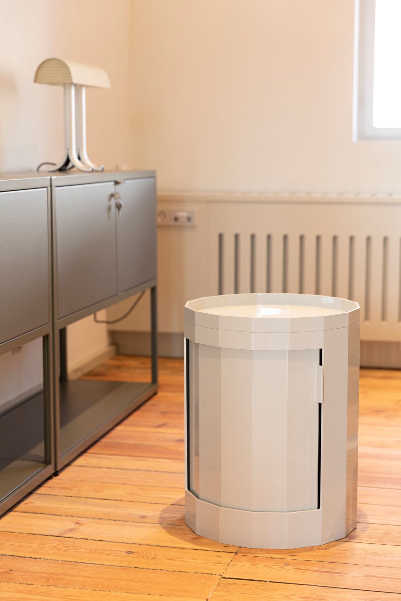 Rundt bord med en hvid lampeskærm og en hvid radiator bagved i en interiør med lyse vægge og trægulv