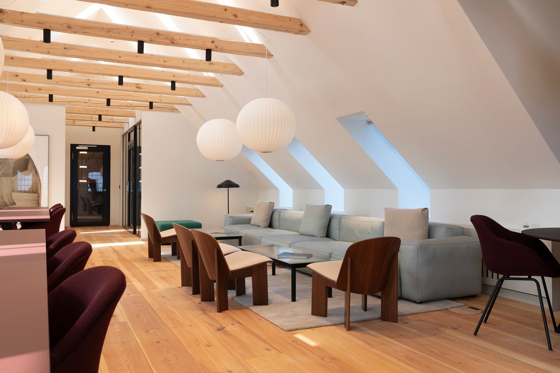 Loftsværelse med lyse loftsbjælker og lysekroner. En stor sofa med putter og et bord med en bøjet lampe. Et skarpt gulv og