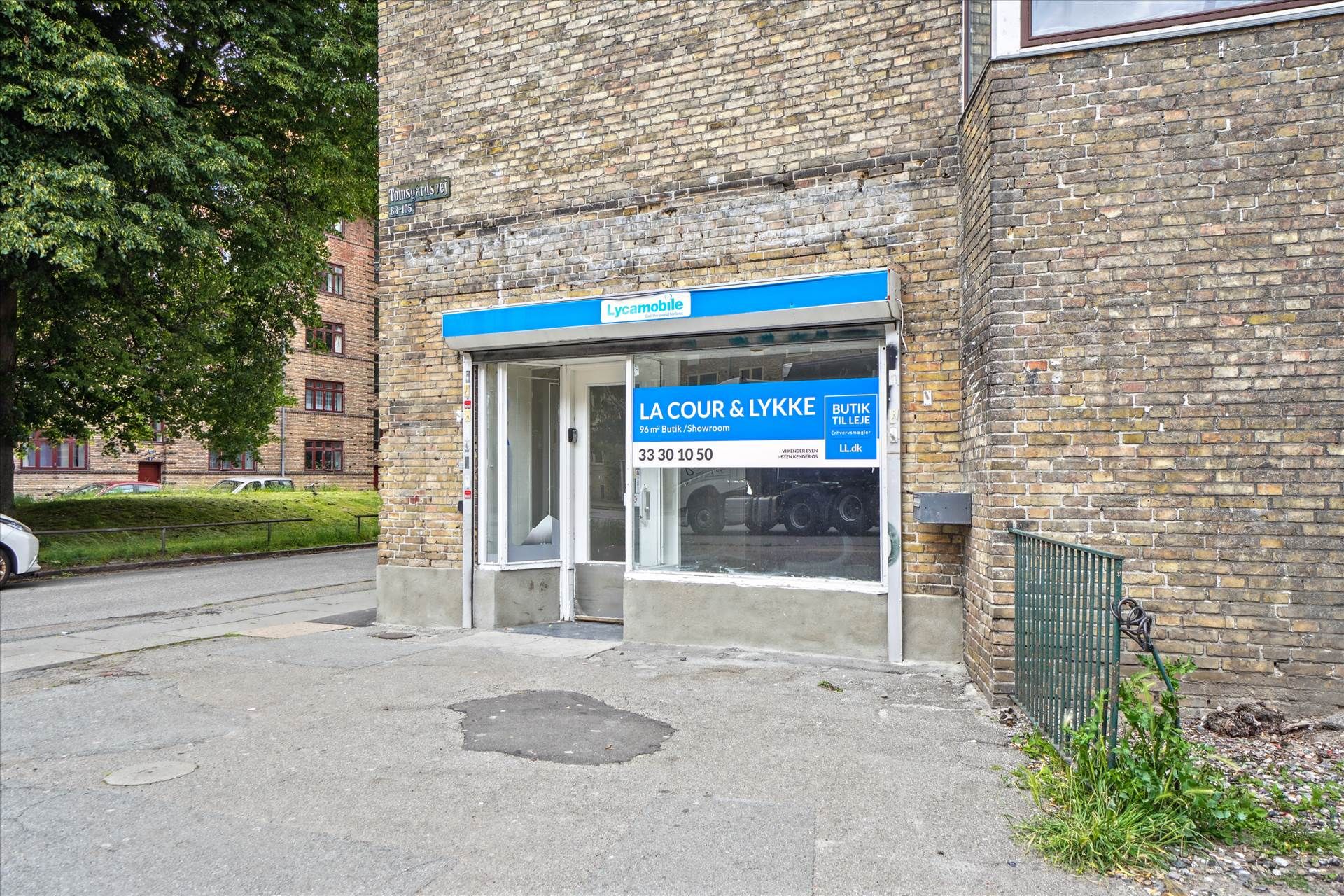Oplysningsskilt for erhvervslokaler i glasdør og med reklamebanner for La Cour & Lykke. Kædebrolægget og vejside med grønne