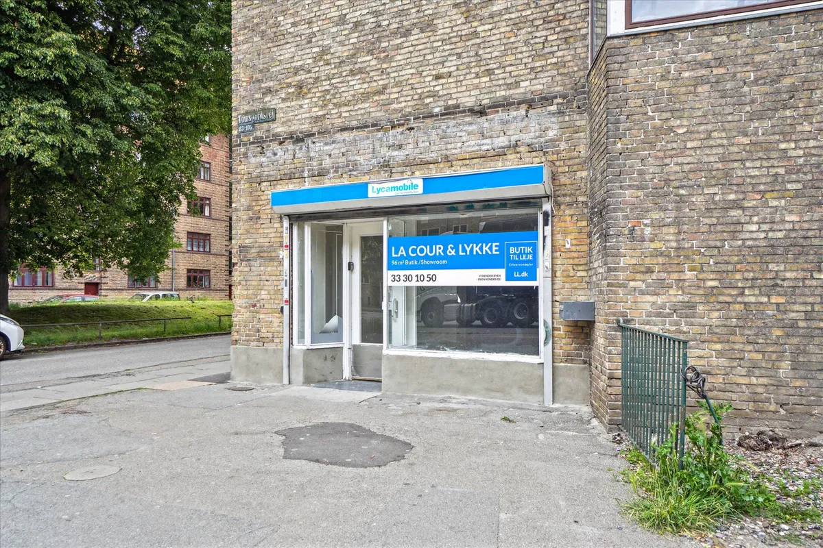 Oplysningsskilt for erhvervslokaler i glasdør og med reklamebanner for La Cour & Lykke. Kædebrolægget og vejside med grønne