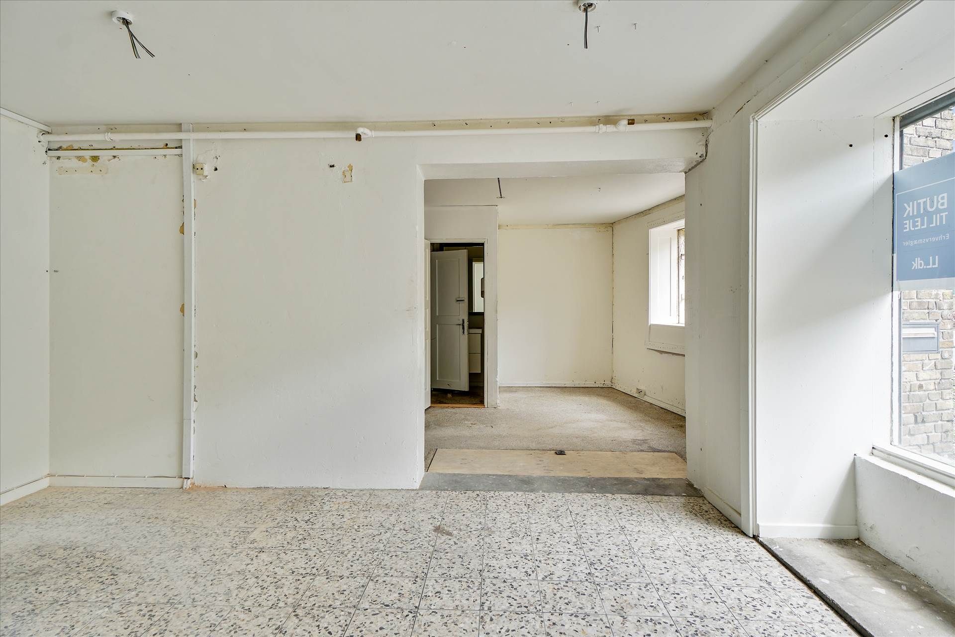 En tom og ren erhvervslokal med lyse vægge, højt til loftet og spidse hjørner samt et vinduesparti med et hvidt skilt og en