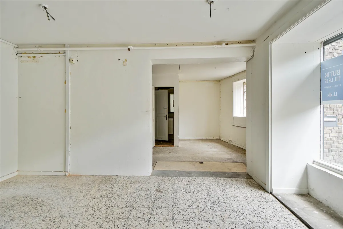 En tom og ren erhvervslokal med lyse vægge, højt til loftet og spidse hjørner samt et vinduesparti med et hvidt skilt og en