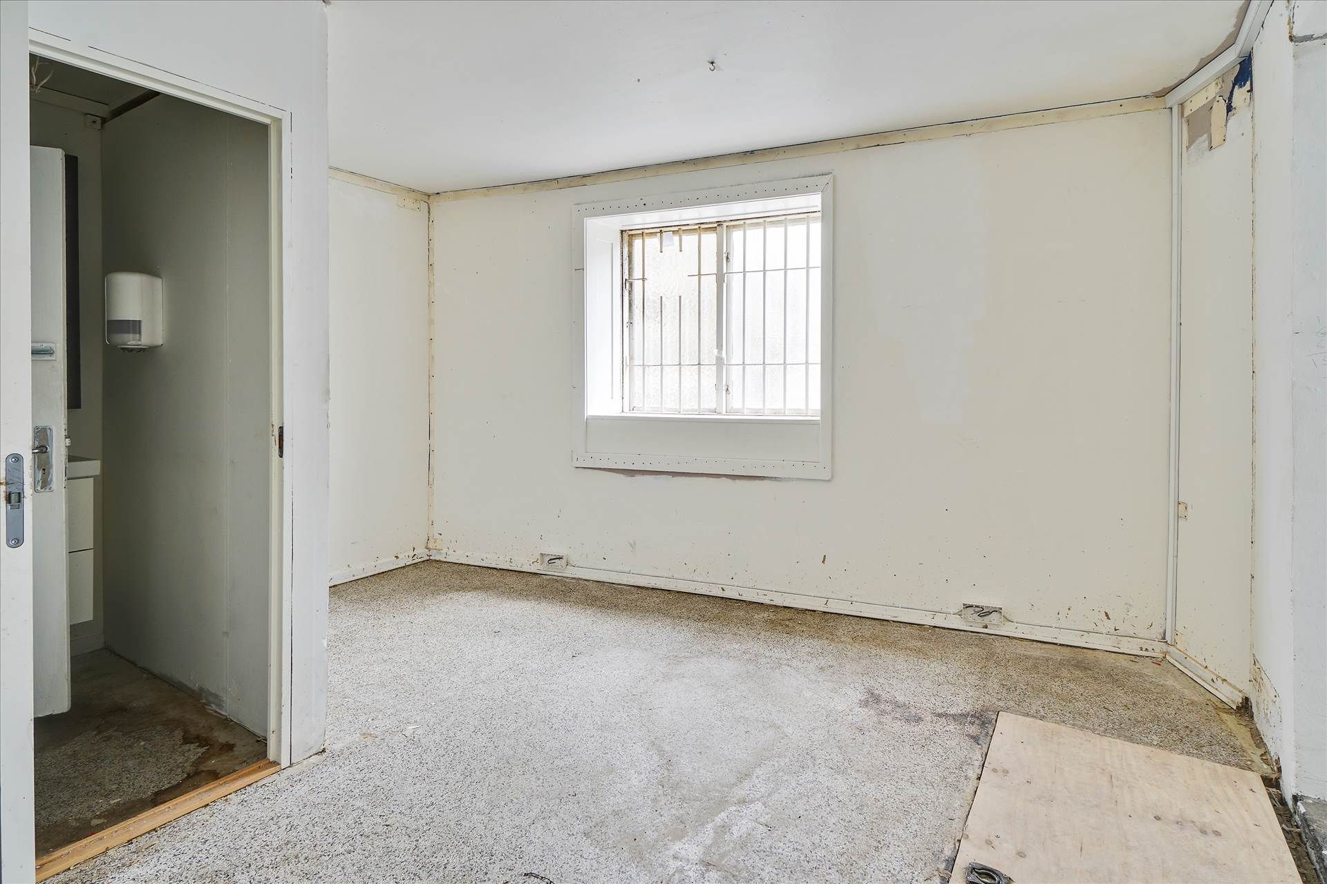 Rum med hvid maling og hvidt loft, vinduesruder med metalgitter og gulv med forskidte pletter og spor