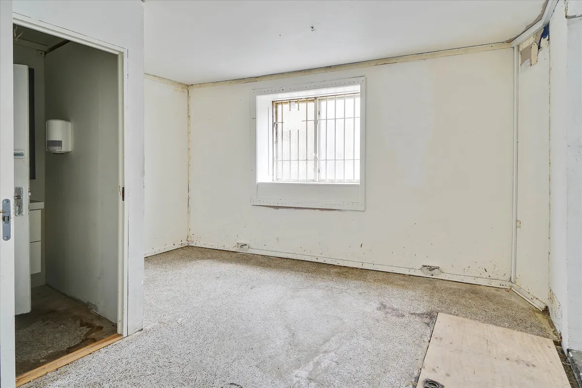 Rum med hvid maling og hvidt loft, vinduesruder med metalgitter og gulv med forskidte pletter og spor