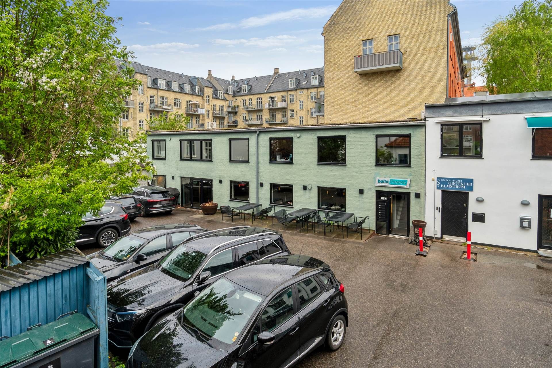 Parkeringsplads med fem sorte biler foran et grønt og hvidt bygningskompleks med teksten Baltic Finance og Advokatfirmaet