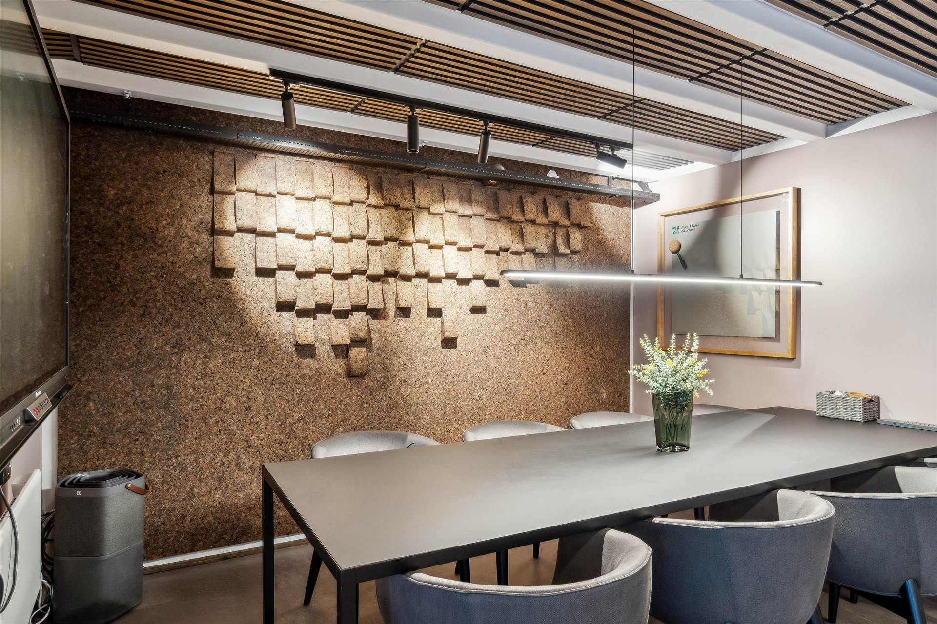 Langt bord med stole, vase med blomster, og en væg med mønstret kork i et moderne kontor med lysegul baggrundsvæg