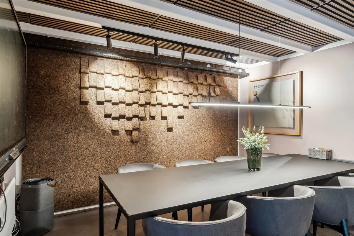 Langt bord med stole, vase med blomster, og en væg med mønstret kork i et moderne kontor med lysegul baggrundsvæg