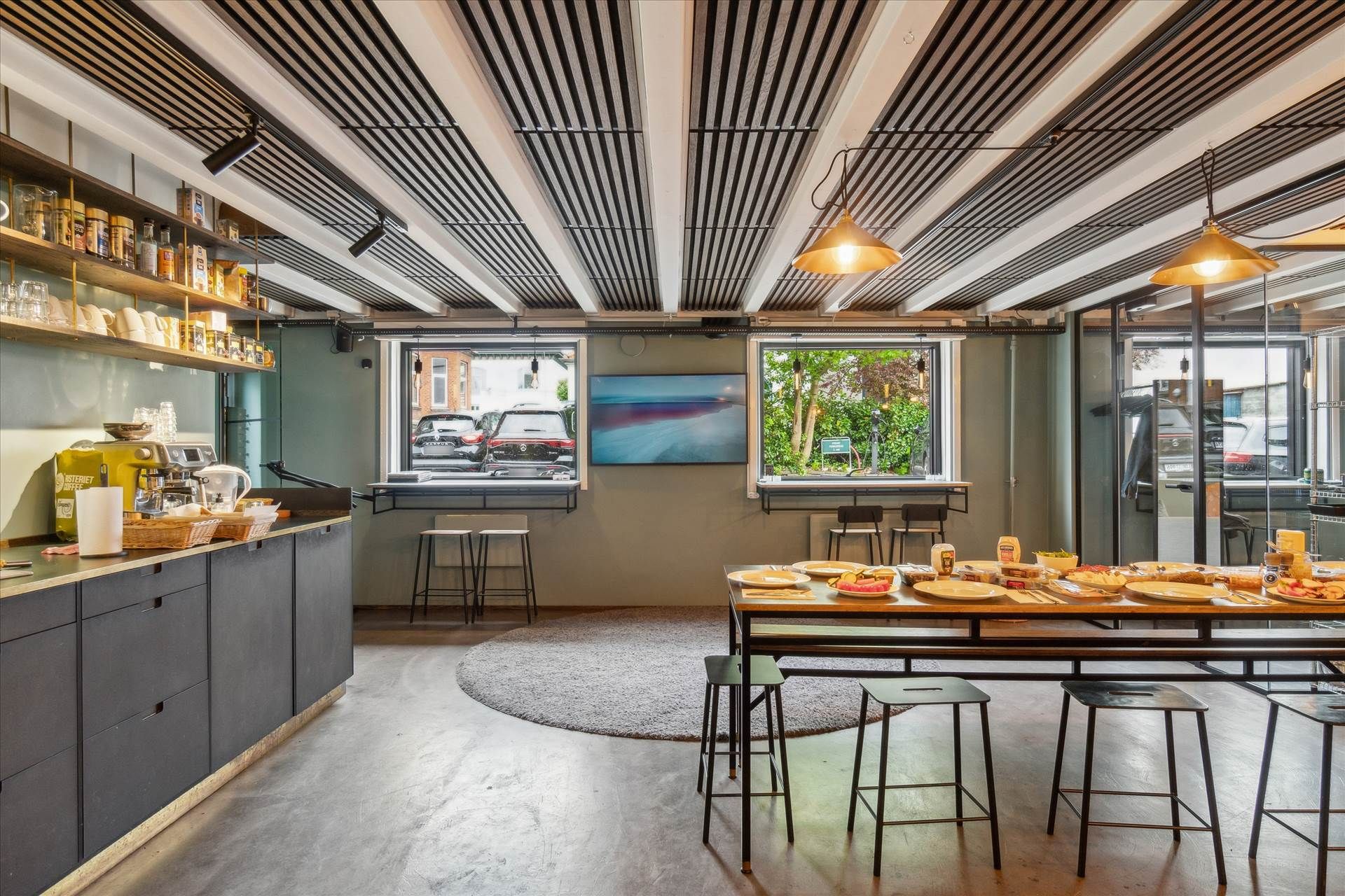 Hyggeligt køkken med lyse loftsbjælker og lysekroner, køkkenudstyr og retter på bordet, gulvtæppe og stole, vindue med