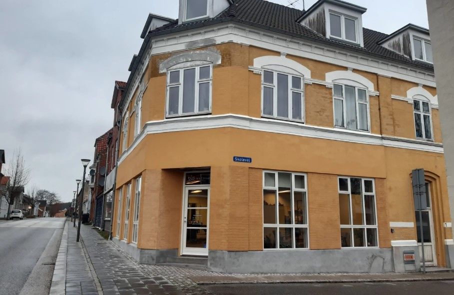 Gult og hvidt hus med store udstillingsvinduer og tekst "Skolevej" på facaden, to søjler og skrapelister langs gaden