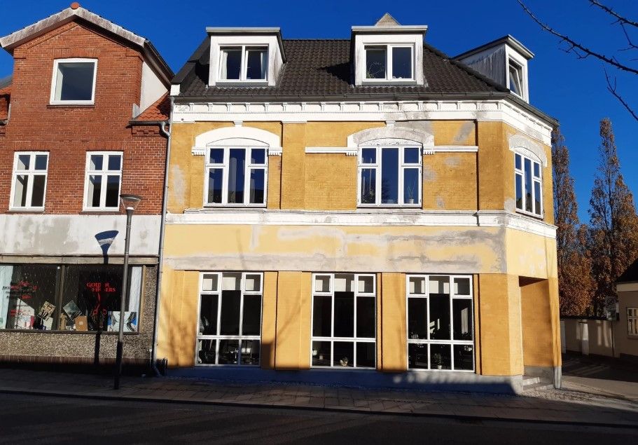 Gul bygning med udstillingsvinduer og to etager, opført i gul og hvid puds. Træstammer og trægrene synlige mod højre
