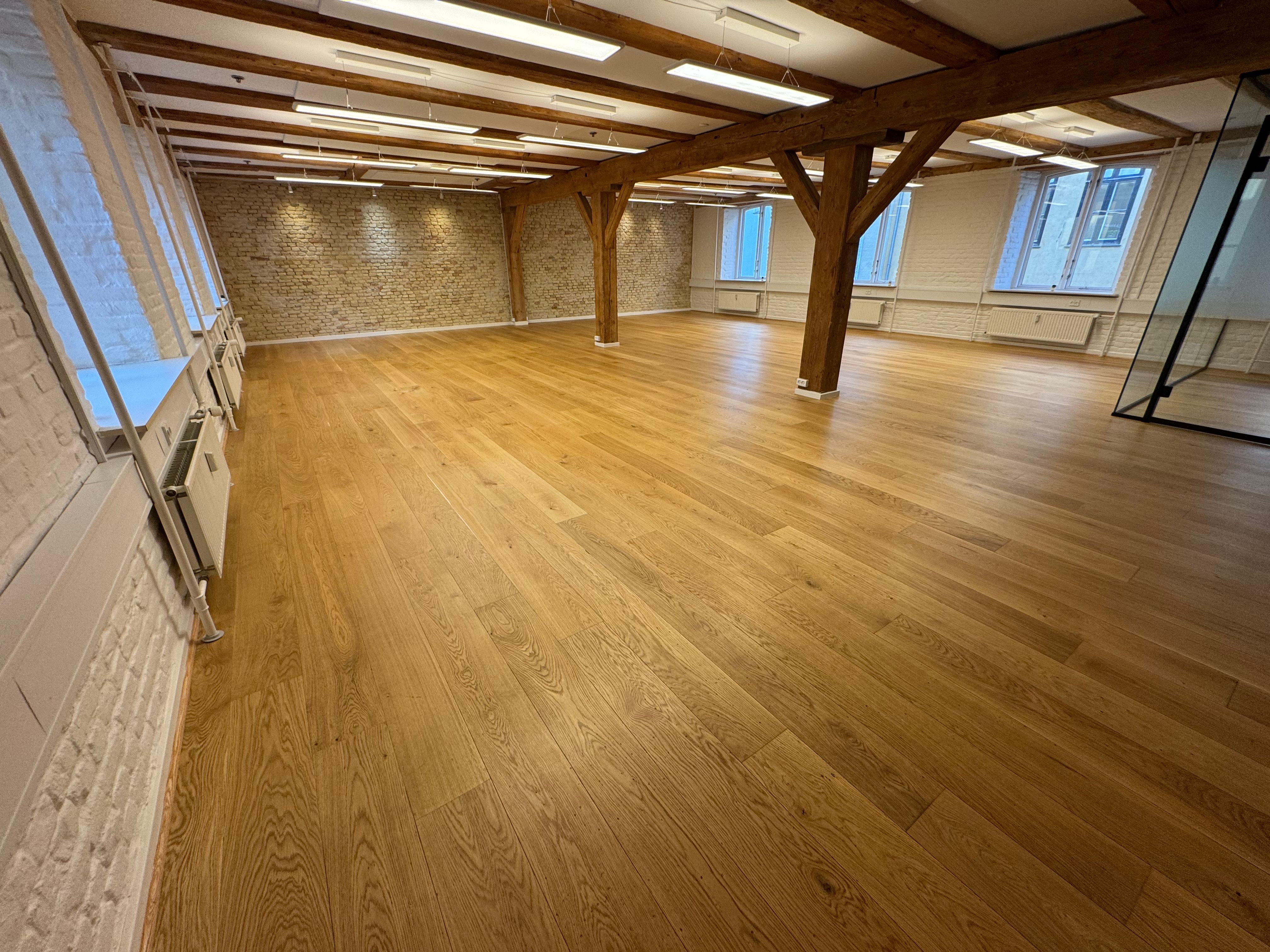 En stor, tom rum med højloft og lyse trægulve. Rækker af lysepunkter og hvide vægge. Træbjælker, der støtter loftet. Vinduer