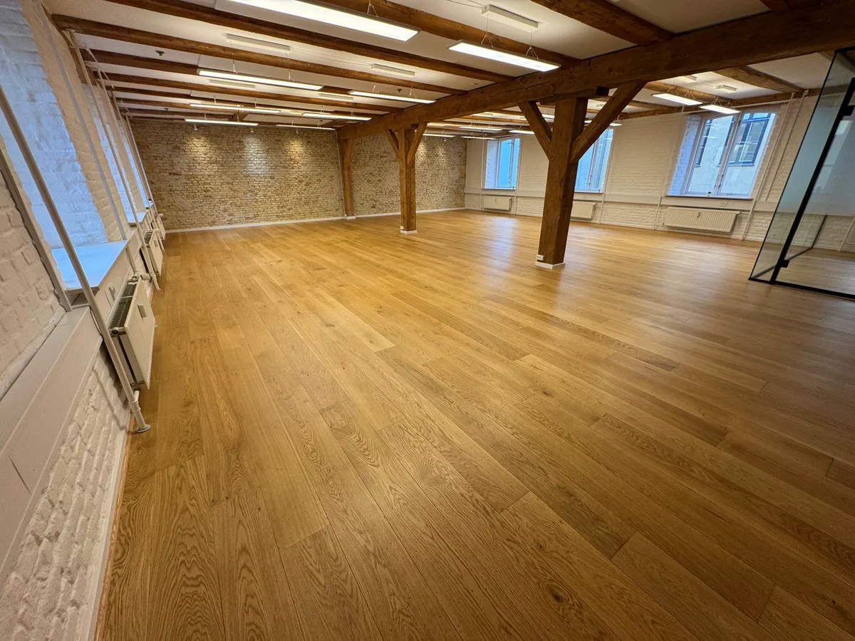 En stor, tom rum med højloft og lyse trægulve. Rækker af lysepunkter og hvide vægge. Træbjælker, der støtter loftet. Vinduer