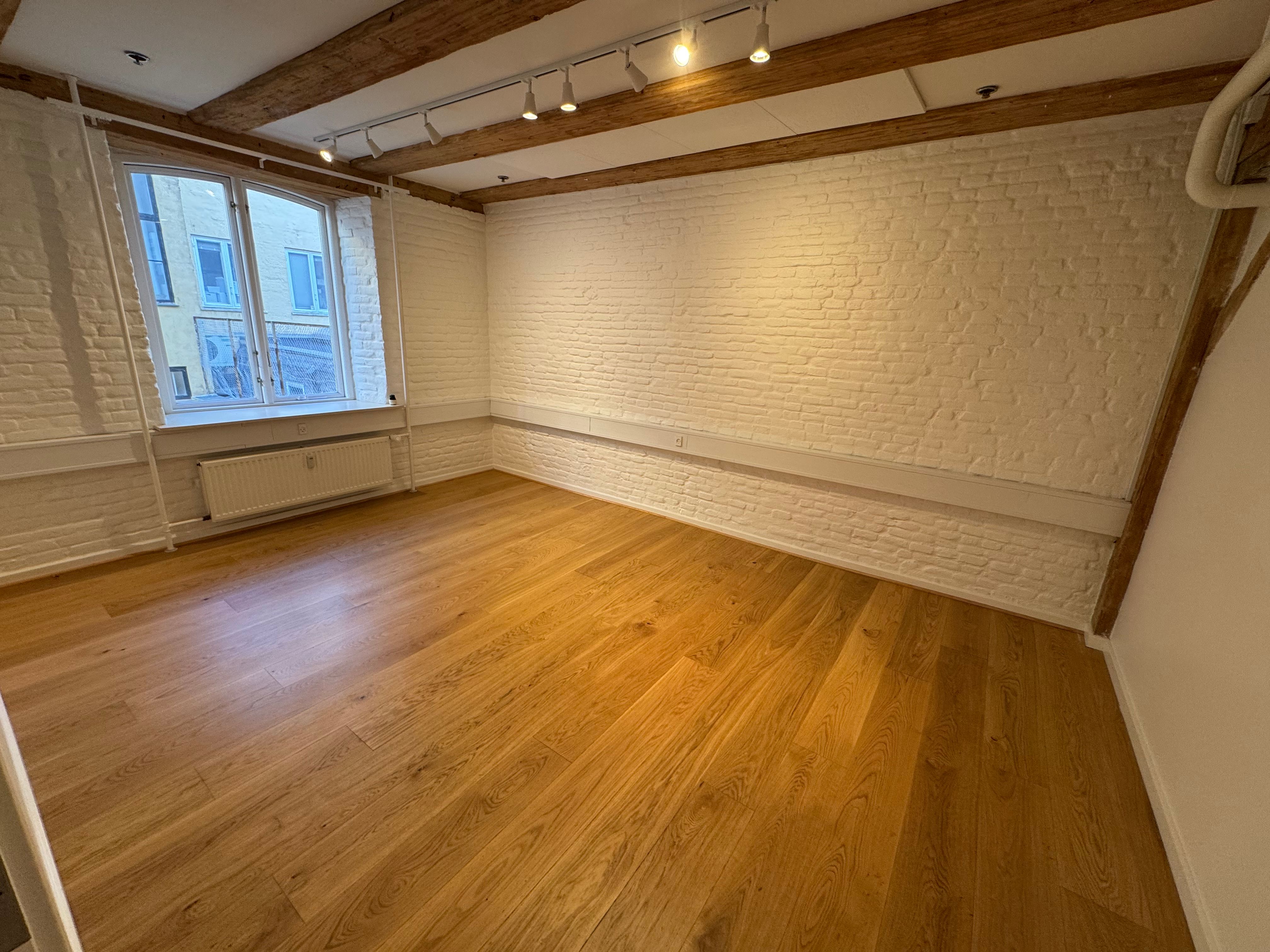 En stor tom rum med højt loft og lysekroner. Trægulv og hvide vægge. Et vindue med udsigt til bygning.