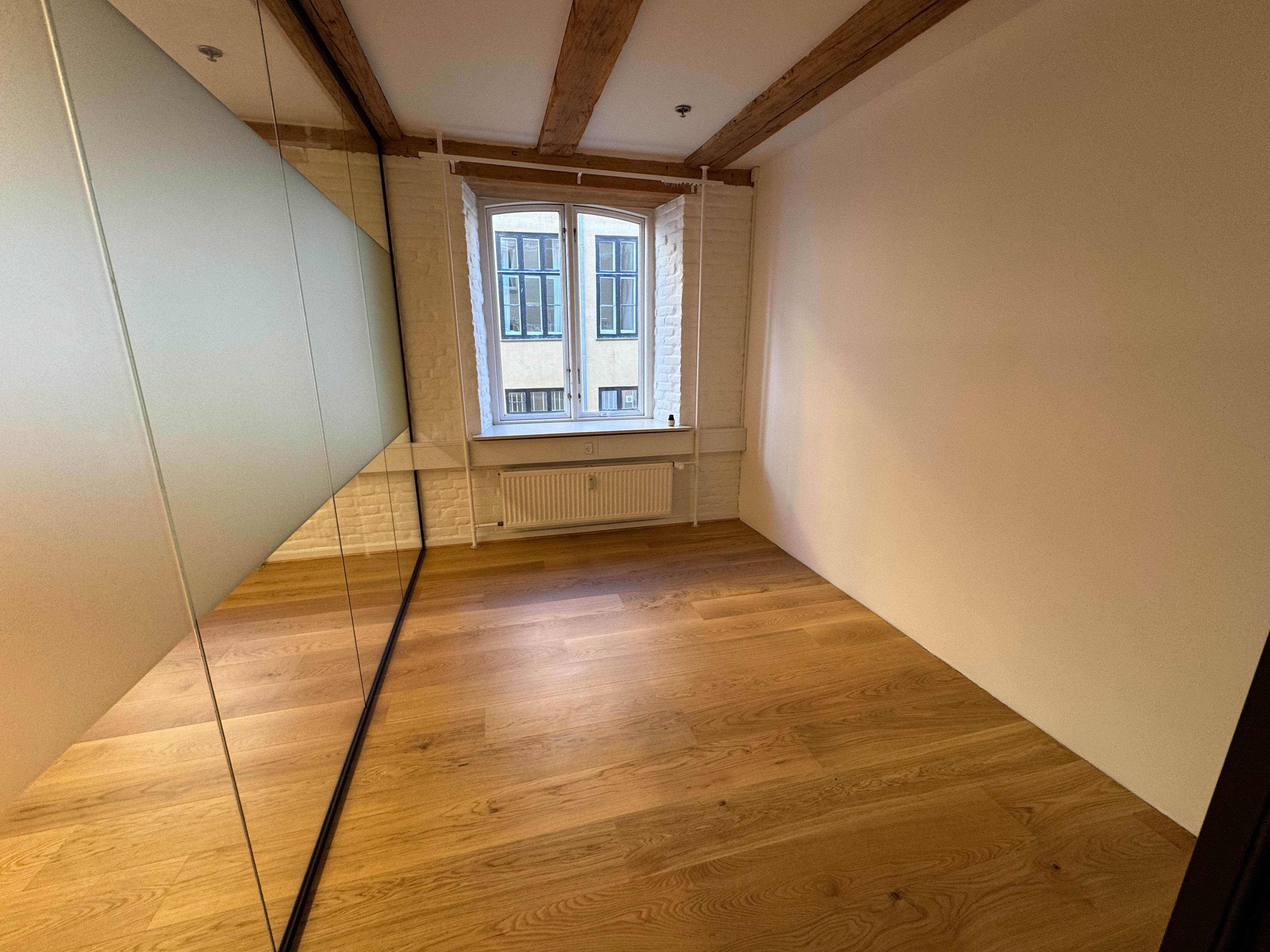 En moderne og minimalistisk stue med blanke gulve, hvide vægge og træbjælker i loftet. En stor spejl og et vindue med